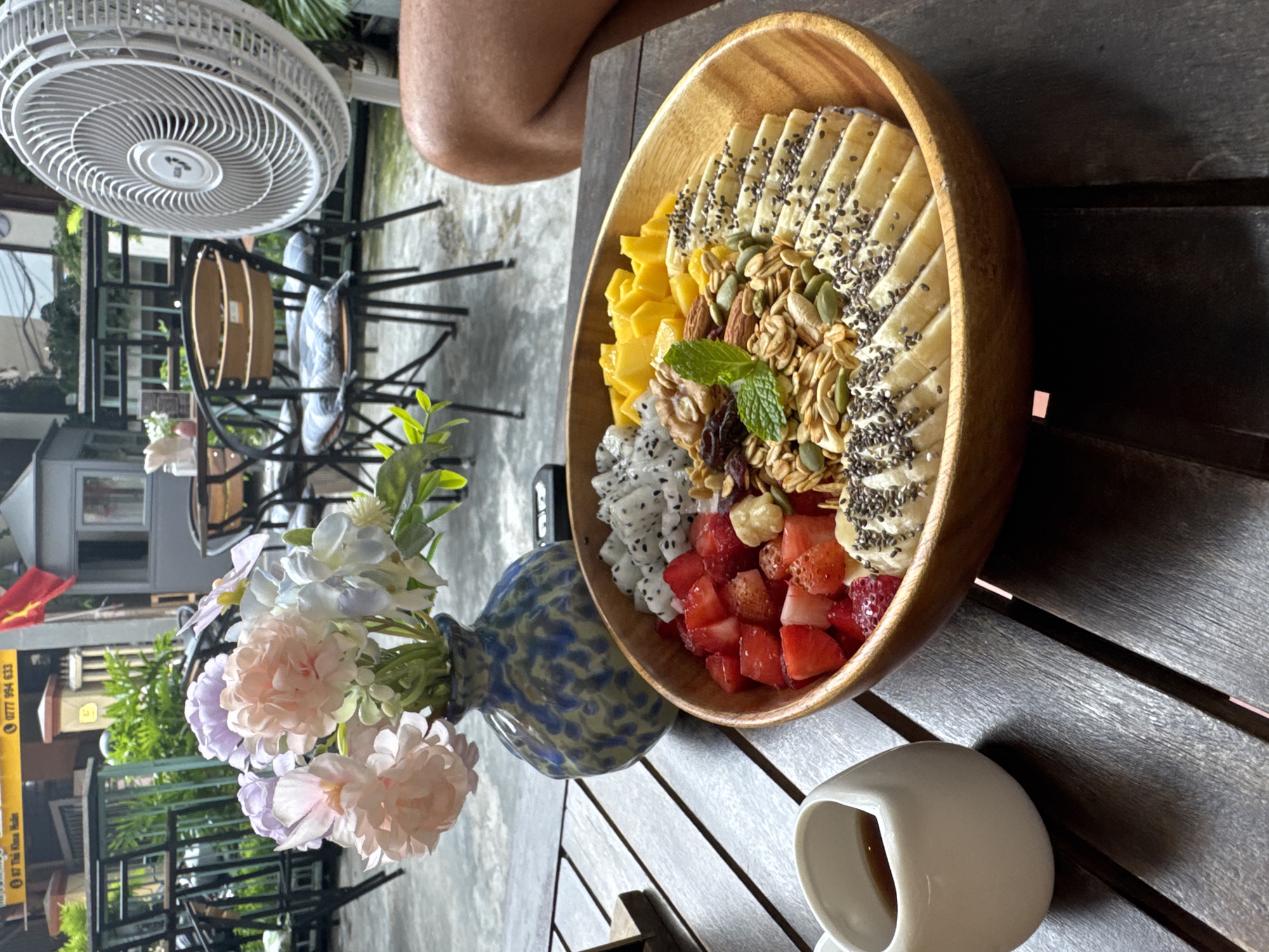 A wooden table in Đà Nẵng, Vietnam, is set with a wooden bowl containing sliced bananas, mango, dragon fruit, and strawberries, topped with granola and a sprig of mint. A white mug sits beside the bowl. A blue vase with light pink and white flowers is also on the table, positioned near the bowl. In the background, visible through the open-air setting, are chairs, tables, a building, and foliage. A fan is visible in the upper left corner. An individual is partially visible in the upper center. The scene appears to be a café or outdoor dining area in Đà Nẵng, Vietnam.