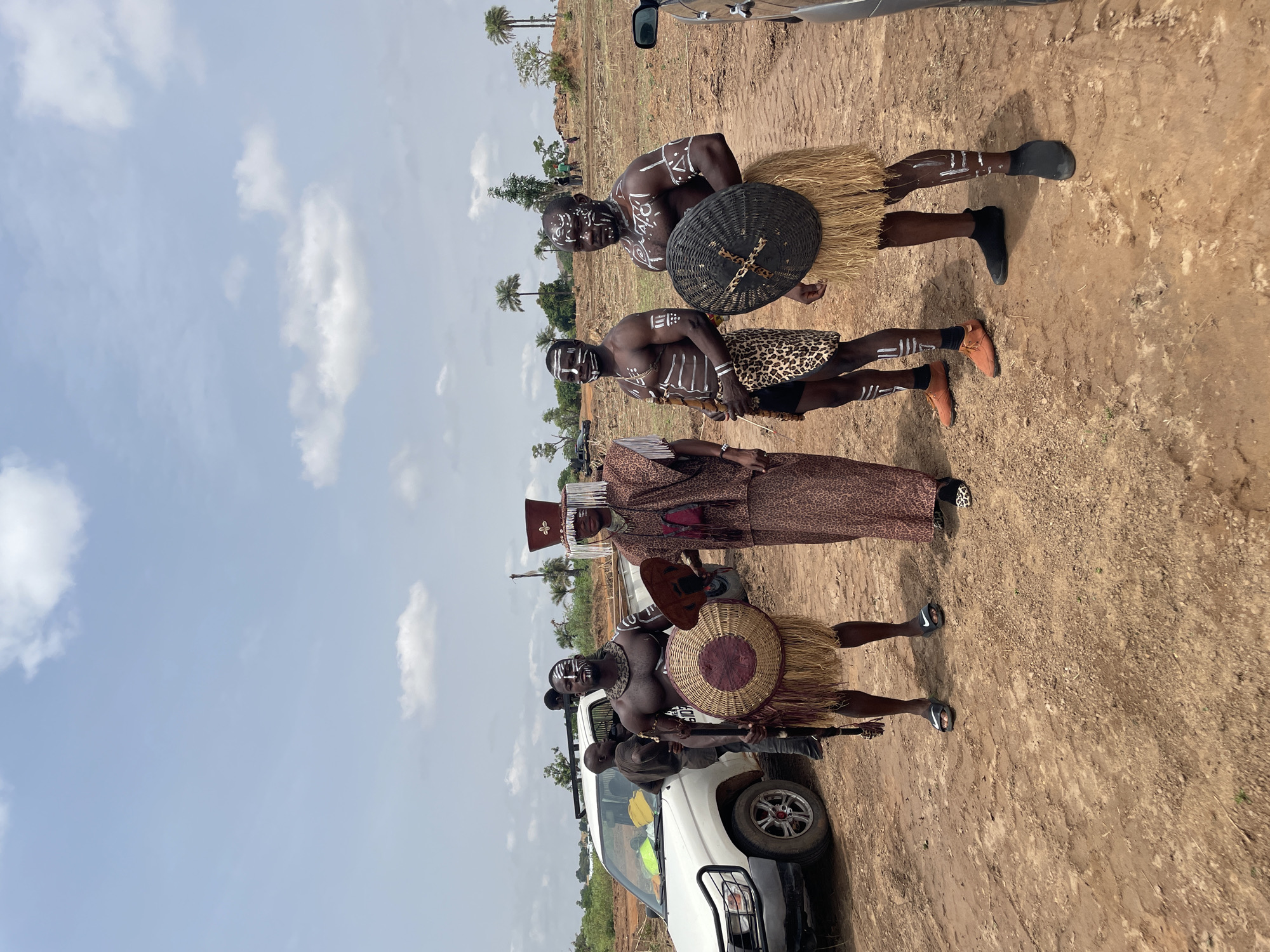 Four African men in traditional attire stand in a dirt field. The men are adorned with white body paint and wear straw skirts or patterned clothing. One man has a headdress. Some carry shields and spears. A white pickup truck is partially visible in the foreground. The location is Akwanga, Nigeria. The sky is light blue with scattered white clouds. Palm trees are visible in the background.