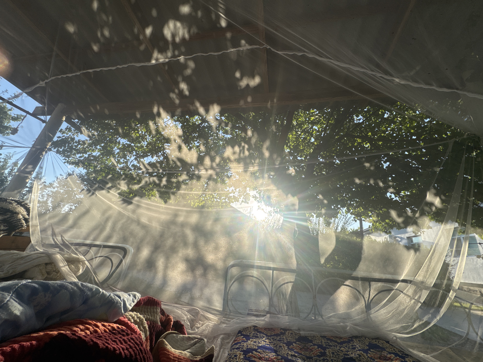 The photograph captures a serene scene inside a screened-in porch or gazebo, likely in the city of Şardara, Kazakhstan. Sunlight streams through the leaves of a nearby tree, casting dappled shadows across the white mesh netting that encloses the space. The netting billows gently, creating a soft, ethereal atmosphere.
Inside, two beds are visible, one covered with a colorful, patterned quilt and the other with a stack of blankets and pillows in muted tones of blue and red. The beds appear to be made of simple metal frames. The overall impression is one of a peaceful, outdoor living space, bathed in the warm glow of the late afternoon sun. The presence of the netting suggests protection from insects, common in outdoor areas, especially during warmer months in regions like Kazakhstan.