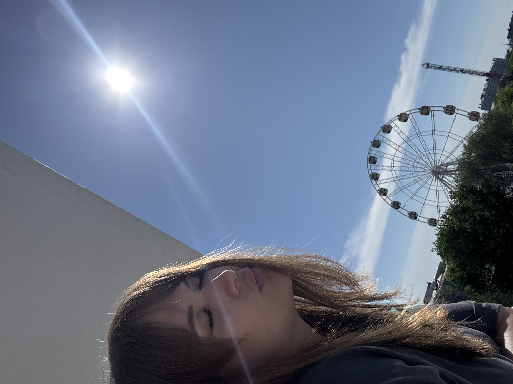 The image is taken from a low angle, looking up at a fairground attraction and the sky. A person with brown hair and closed eyes occupies the lower foreground. In the background, a Ferris wheel is visible to the right, with a tall tower ride partially visible behind it, along with some green trees. The sky is blue with a bright sun to the left and a few wispy clouds near the Ferris wheel. A white structure is visible on the left side of the image. It is likely that this scene is in the city of Yevpatoriya, Ukraine due to the fairground rides.