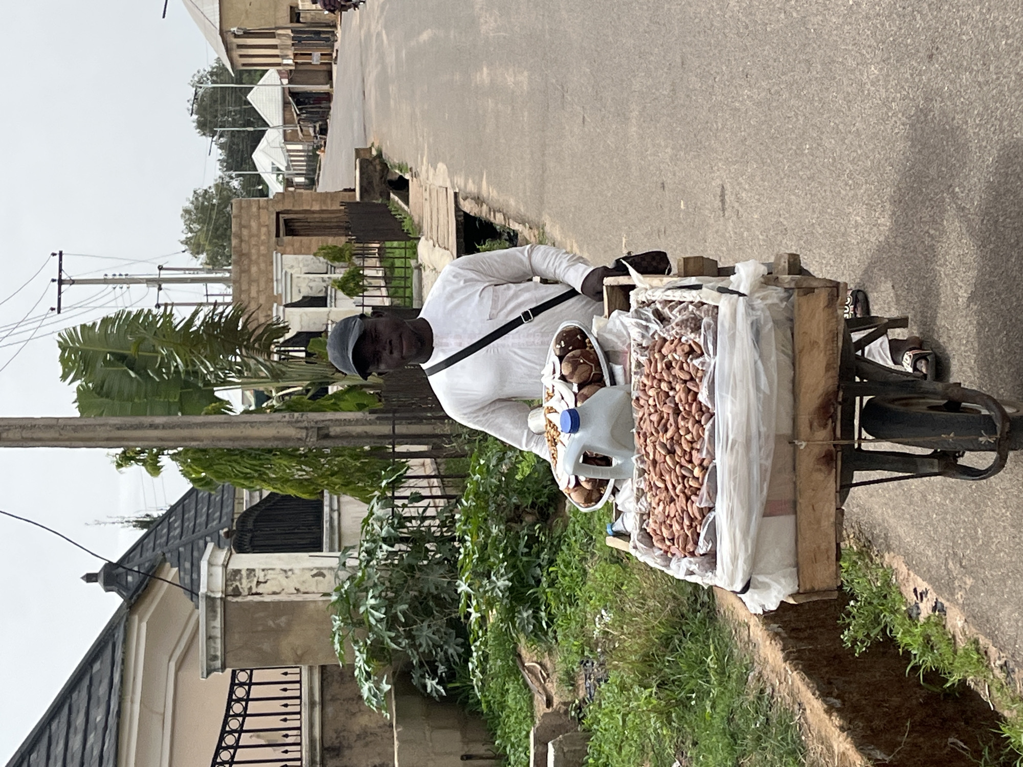 A man wearing a white long-sleeved shirt and a dark cap stands next to a tricycle on a road in Barakin Akawo, Nigeria. The tricycle has a wooden frame holding a large quantity of what appear to be peanuts or other nuts wrapped in clear plastic. A white plastic container filled with round objects, possibly baked goods or more nuts, sits near the packaged nuts. The man has a black strap across his chest.
The background includes residential buildings, utility poles, and greenery. The road is paved and there's a drainage ditch running alongside it. The lighting suggests it's daytime, likely under overcast conditions.