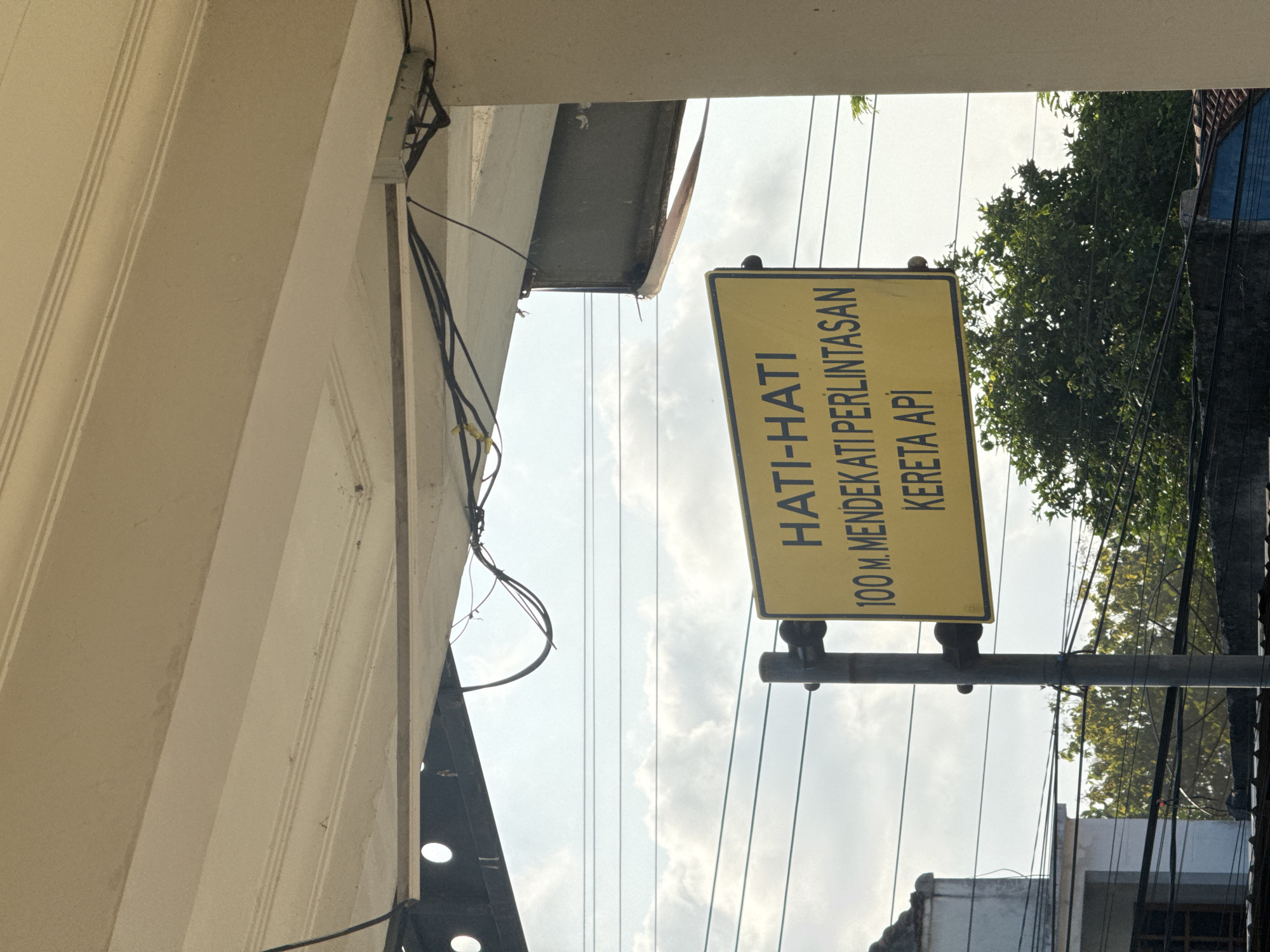 The image captures a close-up view of a yellow traffic sign in Kedawung, Indonesia. The sign, mounted on a metal pole, displays text in Indonesian: "HATI-HATI 100 M. MENDEKATI PERLINTASAN KERETA API". This translates to "CAUTION 100 M. APPROACHING RAILWAY CROSSING". The sign is positioned under an overhang of a building, with wires and conduits running above it. In the background, power lines crisscross the sky, which is partly cloudy. Trees and rooftops of buildings are visible at the bottom of the frame, suggesting an urban or suburban setting. The overall scene is devoid of people or any ongoing activity.