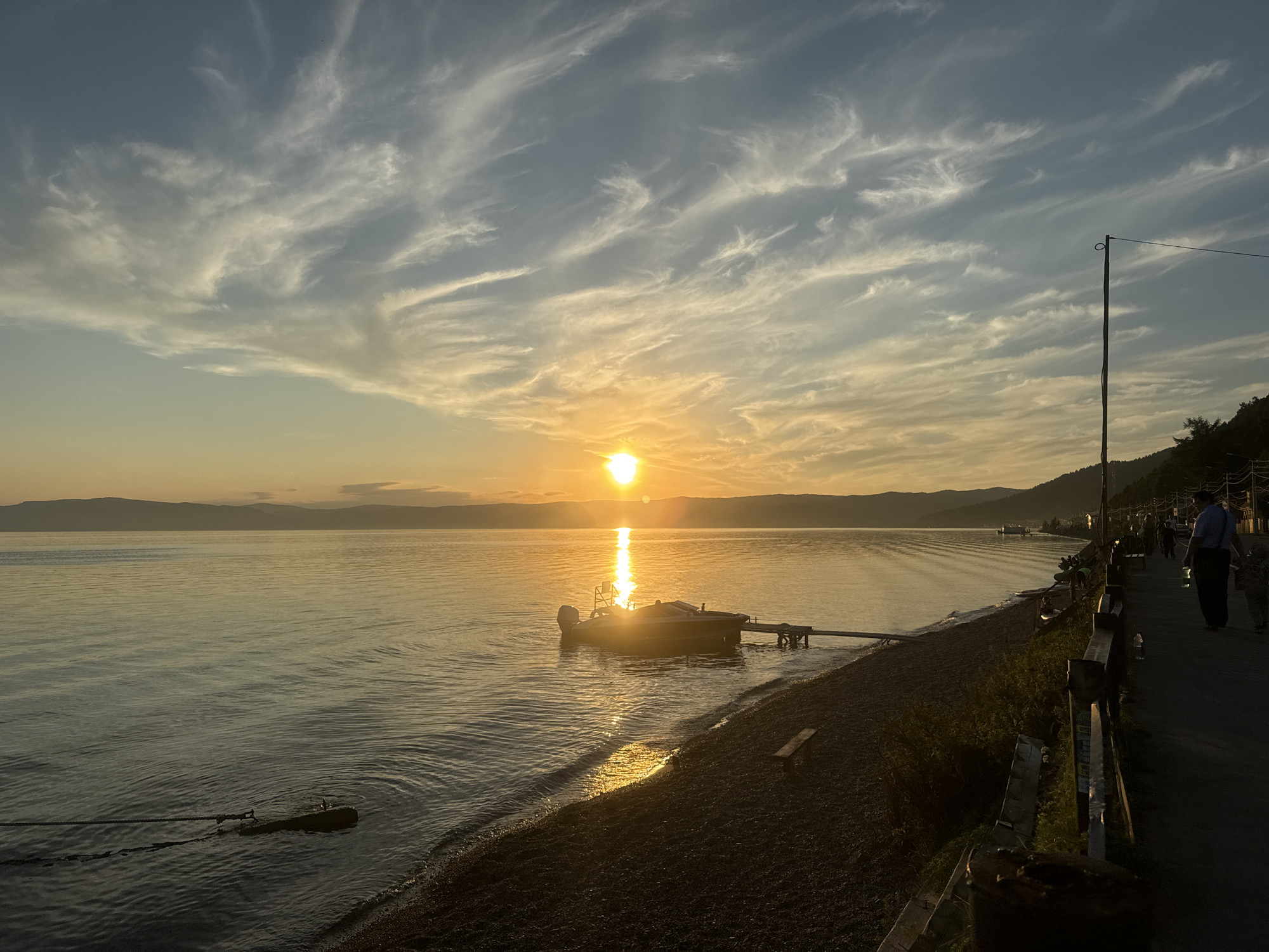 The image depicts a sunset over Lake Baikal in Listvyanka, Russia. The sun is low on the horizon, casting a warm golden glow across the water and illuminating the wispy clouds in the sky. A small motorboat is moored to a wooden jetty, its reflection shimmering on the calm surface of the lake. The shoreline is lined with pebbles and vegetation, and a paved walkway runs alongside it. In the distance, a line of hills forms the backdrop to the scene. A few people are visible walking along the path, silhouetted against the fading light. The overall atmosphere is serene and peaceful, capturing the beauty of a Siberian sunset.
