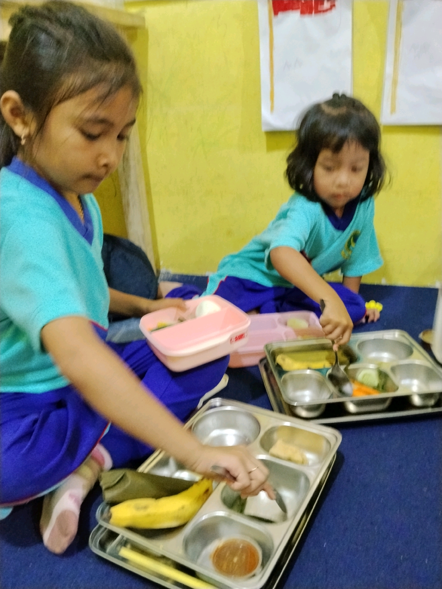 The image depicts two young girls in what appears to be a classroom or common area in Panggungan, Indonesia. Both girls are wearing matching teal t-shirts with blue collars and blue pants. The girl on the left is positioned in the foreground, leaning forward and looking down at a lunch tray. She has dark hair pulled back in a ponytail and is wearing a gold hoop earring in her left ear. Her right arm is extended towards the tray. The girl on the right is in the background, also looking down. She has dark, shoulder-length hair.
In front of the girls are two metal lunch trays with multiple compartments. The tray in the foreground contains a banana, a small container of brown food, and what appears to be some green vegetables and possibly rice. The tray in the background has compartments with food items, including what looks like a rectangular piece of cooked food, sliced cucumbers, and carrots. There is also a pink plastic container with a lid in front of the girl on the left. The floor is covered with a dark blue carpet, and the background wall is painted a bright yellow. Two pieces of white paper with some red markings are pinned to the wall. The overall scene suggests a mealtime activity for young children.