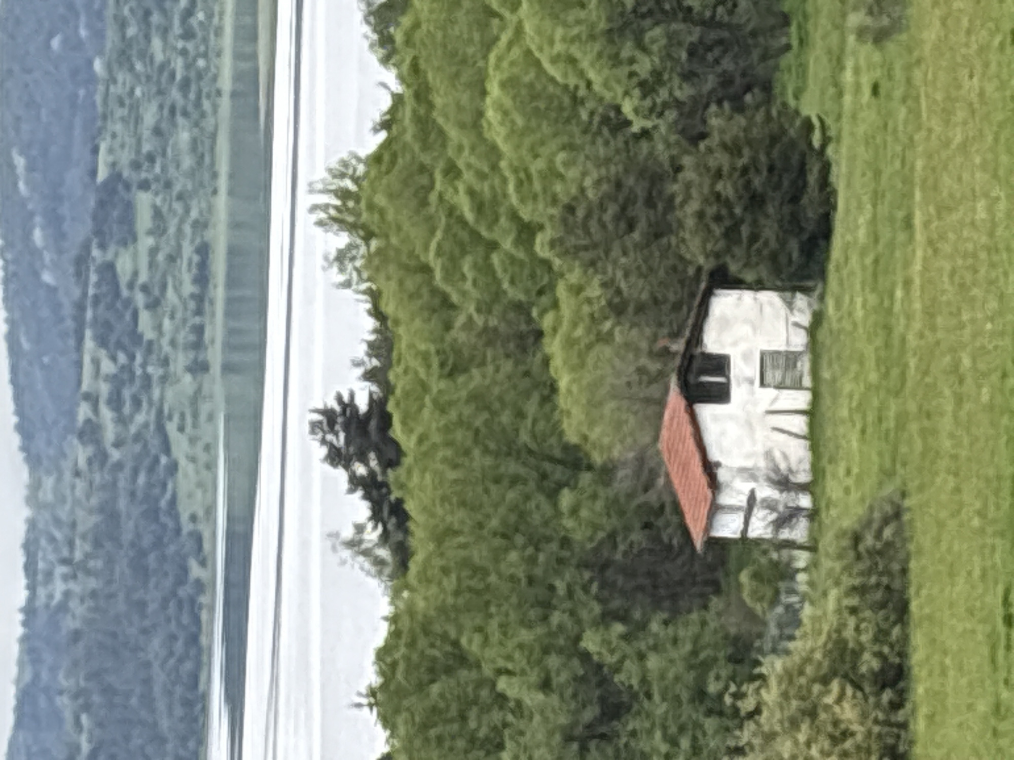 The image depicts a house with a red roof, surrounded by green foliage and a field. In the distance, a body of water, likely a lake, is visible with forested land on the opposite shore. The location is possibly Tsigov chark, Bulgaria, based on the setting and available geographic information. There are no visible people or specific actions taking place. The image primarily captures a landscape featuring a house near a lake surrounded by trees and vegetation.