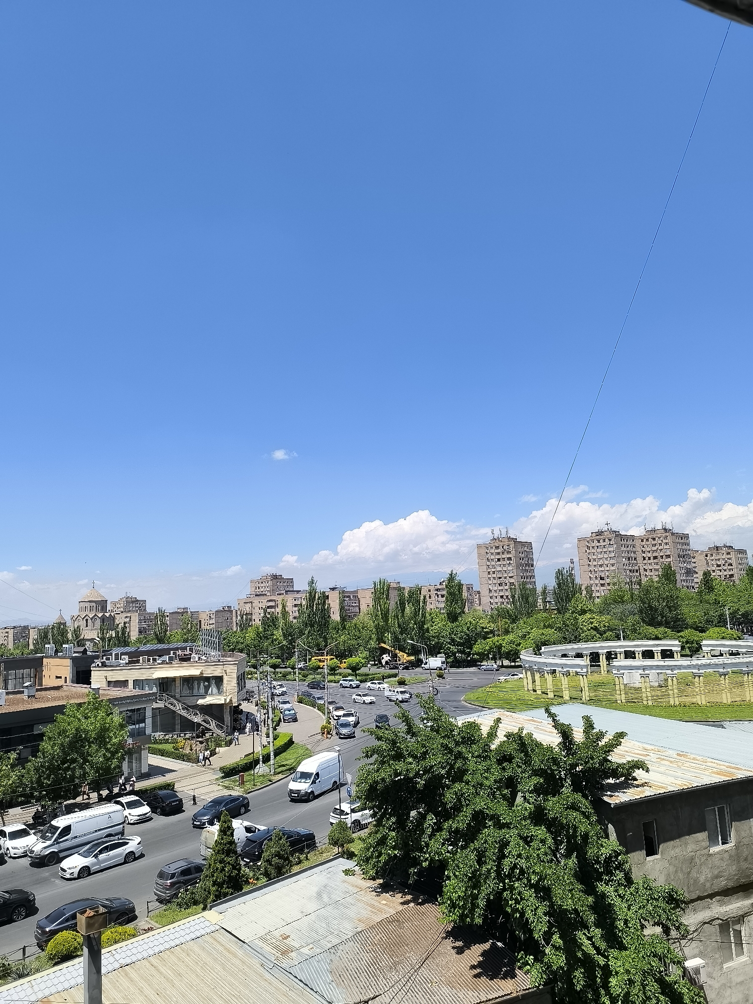 This elevated wide-angle shot captures a vibrant urban scene in Yerevan, Armenia, on a clear, sunny day. The perspective looks down onto a busy street intersection and surrounding city blocks.
In the foreground, the corrugated metal roofs of lower buildings are visible, partially obscured by a large, leafy green tree. Beyond this, a multi-lane road carries a moderate flow of traffic, including various cars, SUVs, and white commercial vans. People can be seen walking on sidewalks and near buildings, indicating typical daily activity.
The mid-ground features a mix of commercial buildings and lush green trees lining the streets. To the left, a distinctive church-like structure with a dome and traditional Armenian architecture stands out amidst other buildings. In the center-right, a unique circular white structure with pillars, possibly a pedestrian bridge or monument, adds a notable architectural element.
The background is dominated by numerous multi-story residential apartment buildings, characteristic of post-Soviet urban planning, interspersed with abundant green trees. Farther in the distance, subtle mountainous terrain is visible under a bright blue sky. The sky itself is expansive and clear, with only a few scattered white cumulus clouds. A thin power line or cable stretches diagonally across the upper right portion of the frame.
The time of day appears to be midday or early afternoon, marked by strong shadows and the bright overhead sun. The weather is fair and sunny. The overall impression is one of a bustling, green city under a wide-open sky. Visible text is minimal; a white van on the left has a stylized "A" logo.