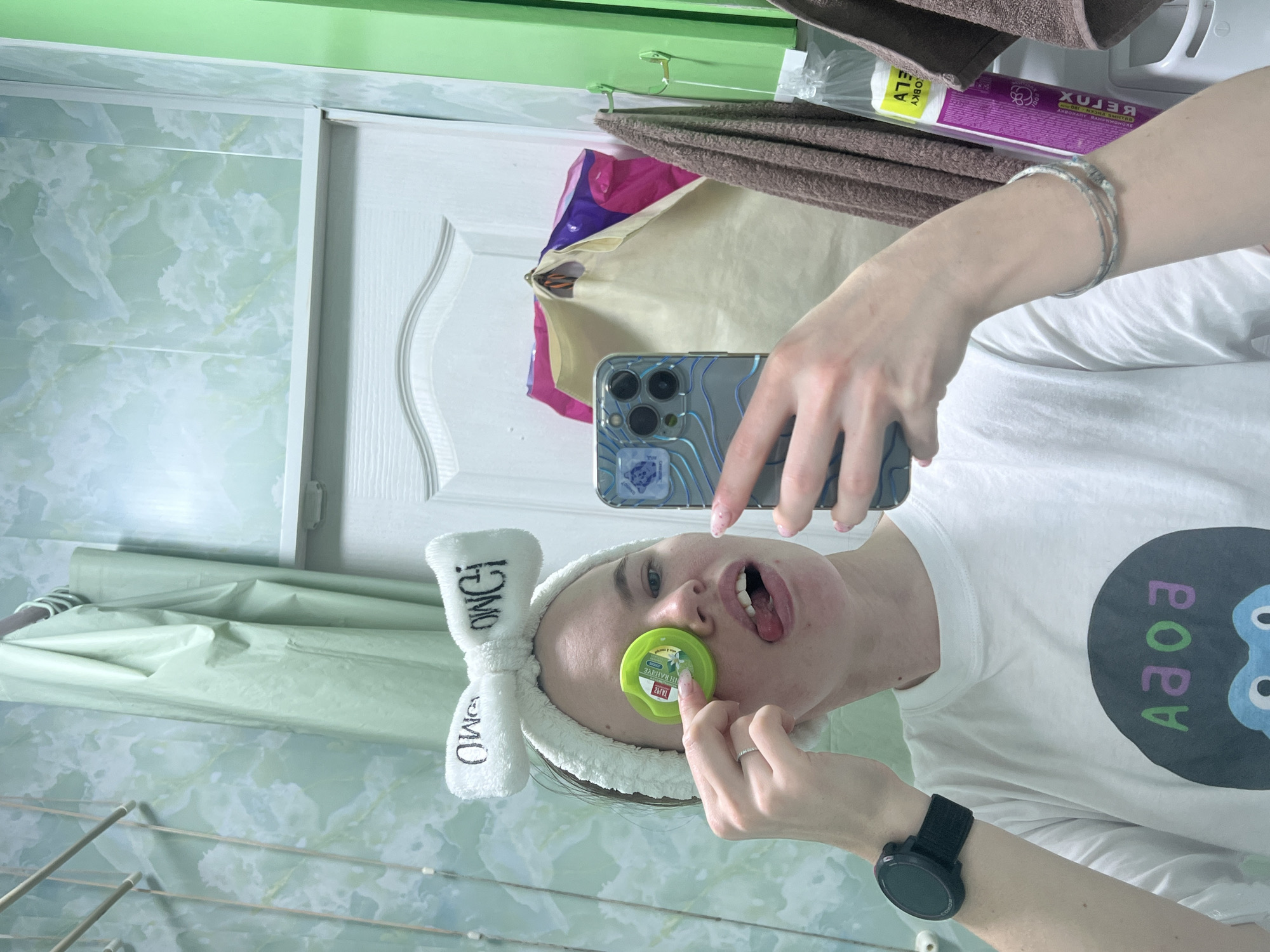 The image depicts a person taking a selfie in what appears to be a bathroom. The person is wearing a white t-shirt with a graphic design, a white headband, and a black smartwatch. They are holding a green container to their face with one hand while using the other hand to take a picture with their phone. The background includes bathroom tiles, a towel rack, and toiletries. Based on textual information, the location is Chaikovsky, Russia.