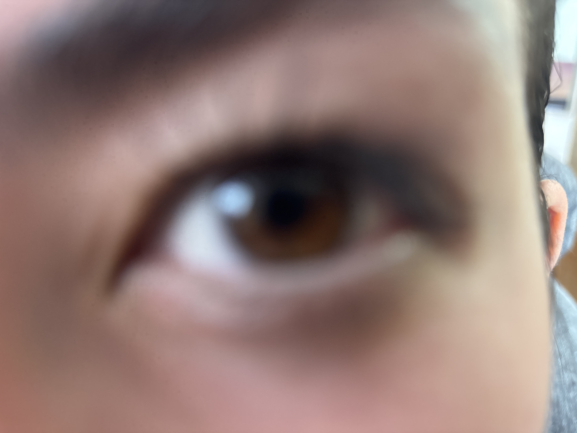 The media file presents an extremely close-up and highly blurred image of a human right eye. The eye's iris is brown, with a distinct light reflection visible, suggesting a light source. The surrounding skin is light to medium-toned, and a dark eyebrow is partially visible along the upper edge of the frame. Towards the far right, a small section of an ear can be seen, adjacent to what appears to be gray fabric, possibly from clothing like a shirt or hoodie. The entire image is significantly out of focus, creating an abstract and soft visual effect. No specific activity or event is depicted beyond this static, intimate view of a facial feature. Due to the blurriness and extreme proximity, details such as the time of day, weather conditions, or specific location cues are not ascertainable. There is no visible text present in the image. The primary focus, despite the lack of sharpness, is the central brown eye.