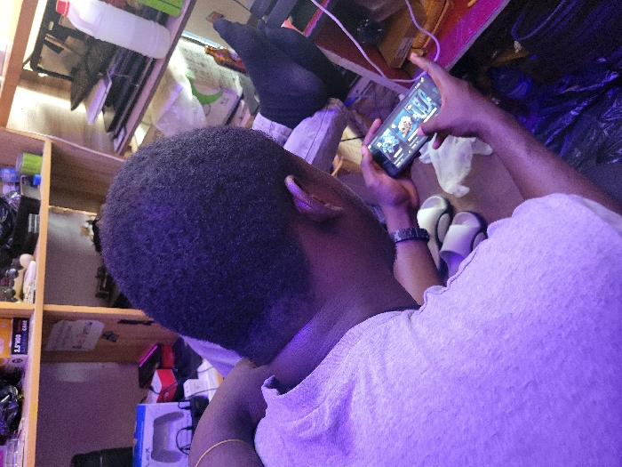 An individual, an adult male with short, dark hair, is depicted from an elevated rear-right perspective in an indoor setting, assumed to be in Makurdi, Nigeria. He wears a light-colored t-shirt, a gold-colored bracelet on his left wrist, and a dark watch on his right wrist. The scene is illuminated by a prominent purple ambient light source. The individual is actively engaged with a black smartphone, held horizontally with both hands, displaying what appears to be a mobile game or application interface. A pair of white and grey/black slip-on sandals and a crumpled white cloth are situated on a surface to his left. In the upper right background, a foot wearing a dark sock is visible, propped up. A light-colored wooden shelving unit occupies the background, containing various items including a box labeled "2.5" HDD CASE," PlayStation 5 controller packaging, several laptops, a white plastic bottle with a red cap, green cylindrical containers, and a brown glass bottle. The immediate floor area features dark plastic materials and visible power cables, including white and pink strands. The environment suggests a cluttered personal space, potentially a room dedicated to electronics or leisure.