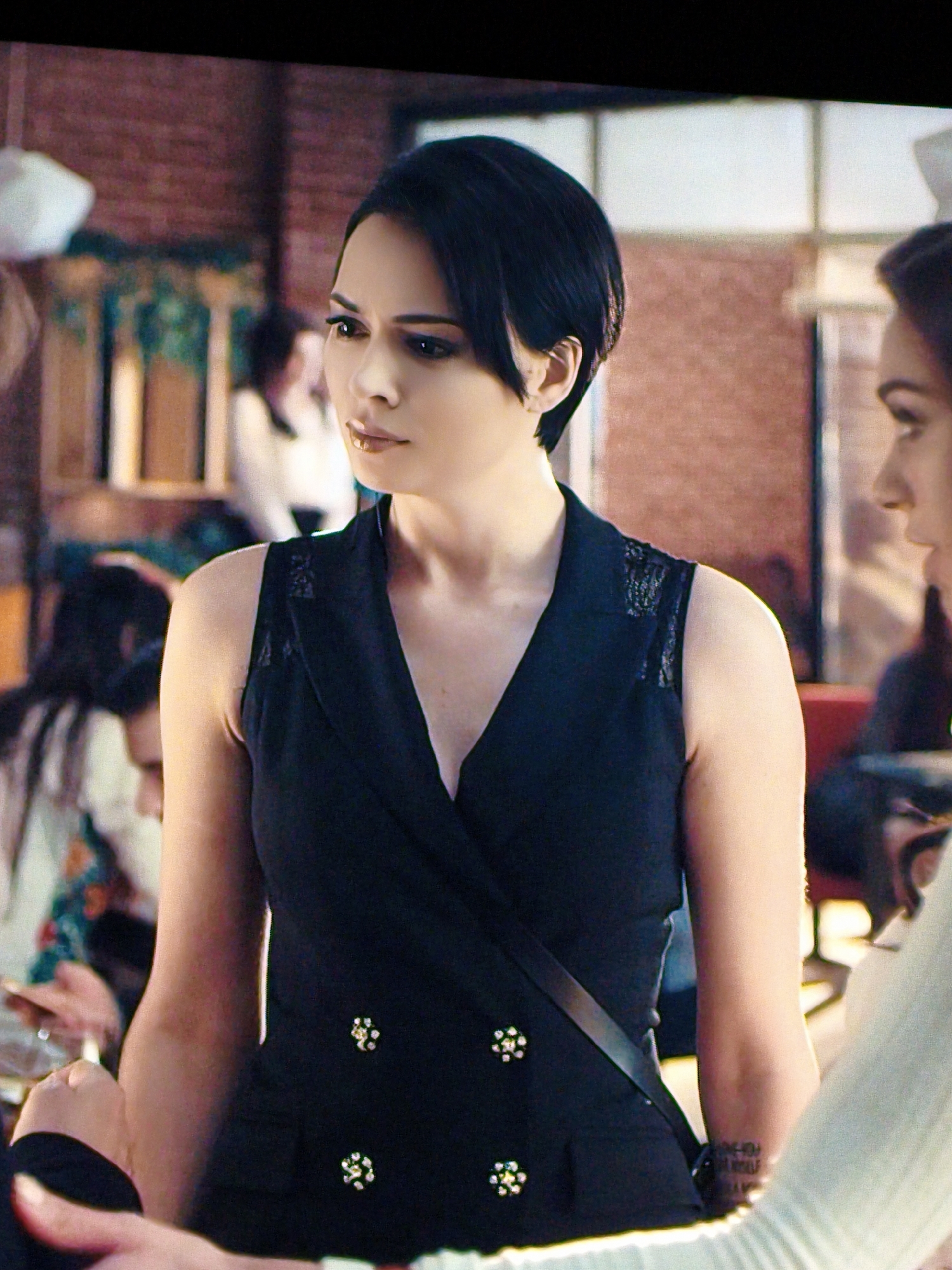 A woman with short black hair is shown from the chest up, looking to her left. She is wearing a black sleeveless dress with a V-neckline and decorative buttons on the front. She has a black strap across her chest. In the background, other people are visible, out of focus, in what appears to be a restaurant or cafe setting. The walls have a brick texture. There are no discernible landmarks or elements that would place the scene specifically in Vladivostok, Russia.