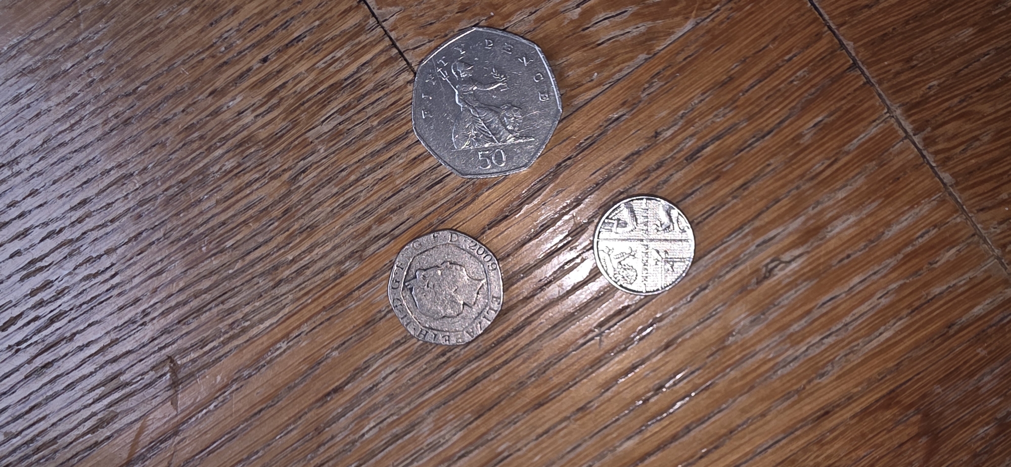 This image shows three British coins on a wooden surface. The coins are a 2009 fifty pence piece, a one pound coin with a shield design, and another coin that is partially obscured. The wooden surface appears to be a floor or tabletop with a prominent wood grain pattern. The lighting is from above, casting slight shadows on the coins. The location of Marum, Netherlands, is not depicted in this image.