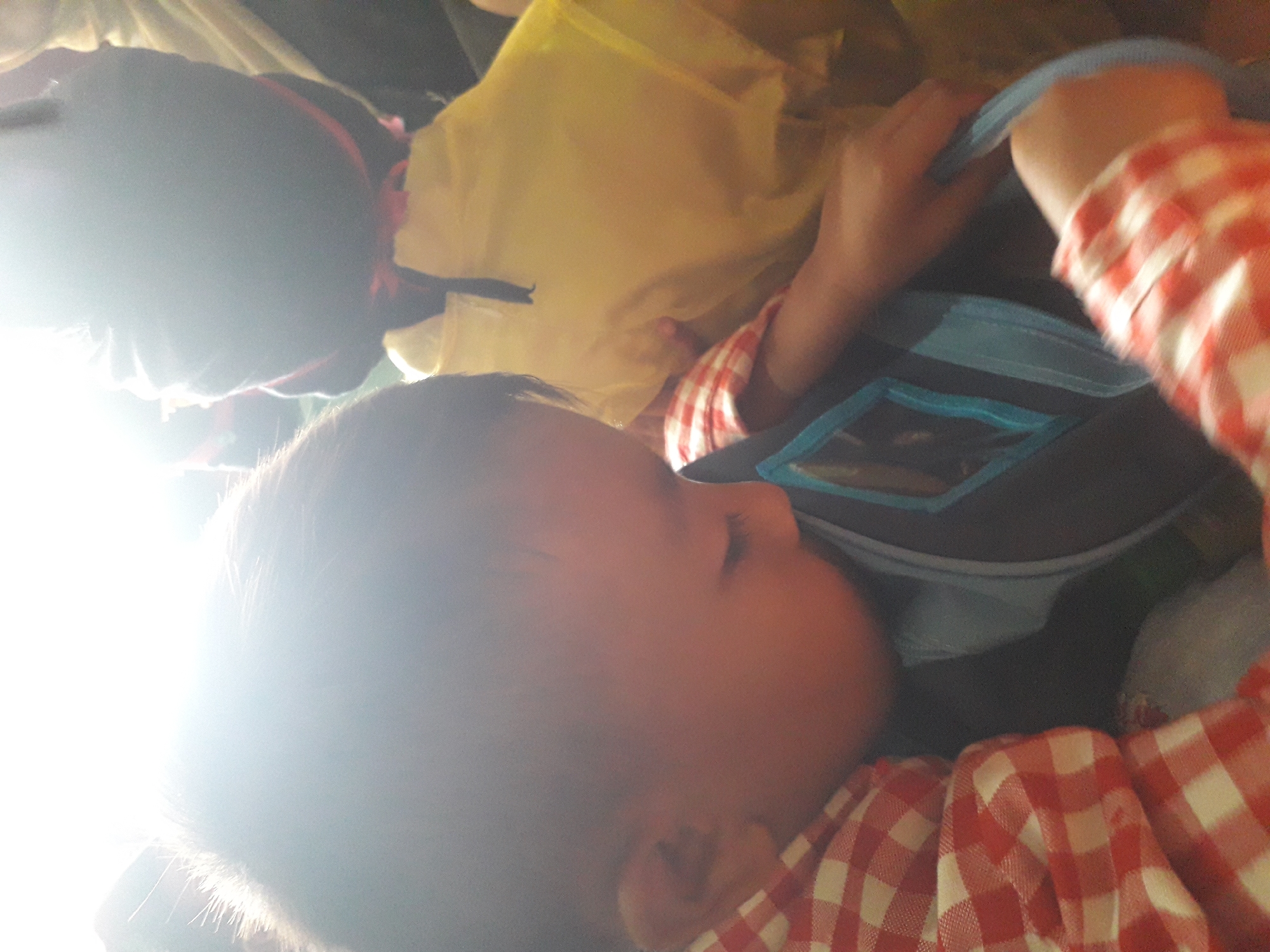 In Cankuang, Indonesia, a close-up shot captures two young children. The child in the foreground, wearing a red and white checkered shirt, leans into a dark backpack with a blue trim, their face obscured by bright light. Another child, seen from behind and wearing a yellow shirt and a dark hat with red ties, is also present. The lighting is harsh and overexposed, creating a strong glare in the upper left of the image. The scene appears to be indoors, possibly in a public transport setting, given the close proximity of the individuals and the limited view of the surroundings.