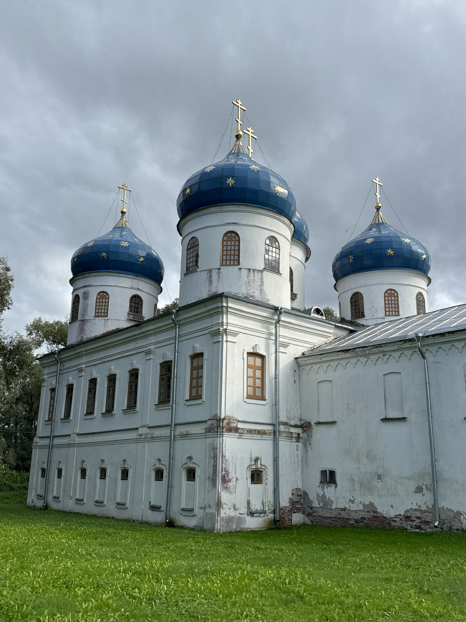 The image shows a large, white, multi-domed Orthodox church with blue domes adorned with gold stars. The church has several windows, some arched and some rectangular. The building shows signs of age with some peeling paint and exposed brickwork at the base of one wall. Green grass covers the ground in front of the church, and a line of trees is visible to the left. The sky is overcast and grey. This is the Church of St. Sophia, located in Veliky Novgorod, Russia. There are no people visible in the image.