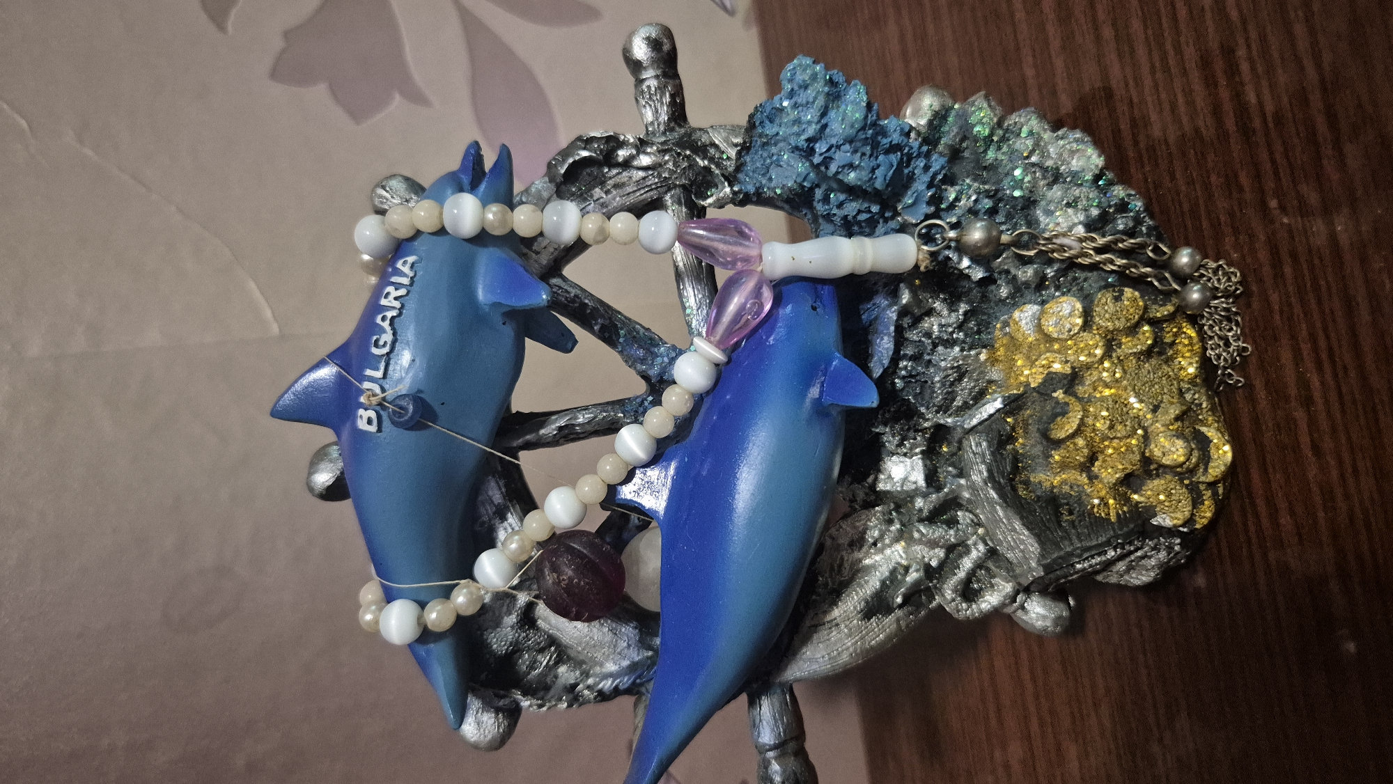 This is a close-up, eye-level, indoor photograph of a souvenir statue featuring two blue dolphins, a ship's wheel, and decorative elements. The statue is positioned on a dark wooden surface, with a mottled brown and grey background. The word "BULGARIA" is inscribed on the upper dolphin. A strand of beads, including pearl-like white beads and a pink pendant, is draped across the dolphins. The base of the statue is decorated with metallic silver accents, gold glitter, and blue, glittery embellishments resembling coral or rocks. The lighting is soft, creating a slight shadow behind the statue. There are no people or animals present in the image. There is no indication of the specific location within the city of Butovo, Bulgaria, or any identifying landmarks.