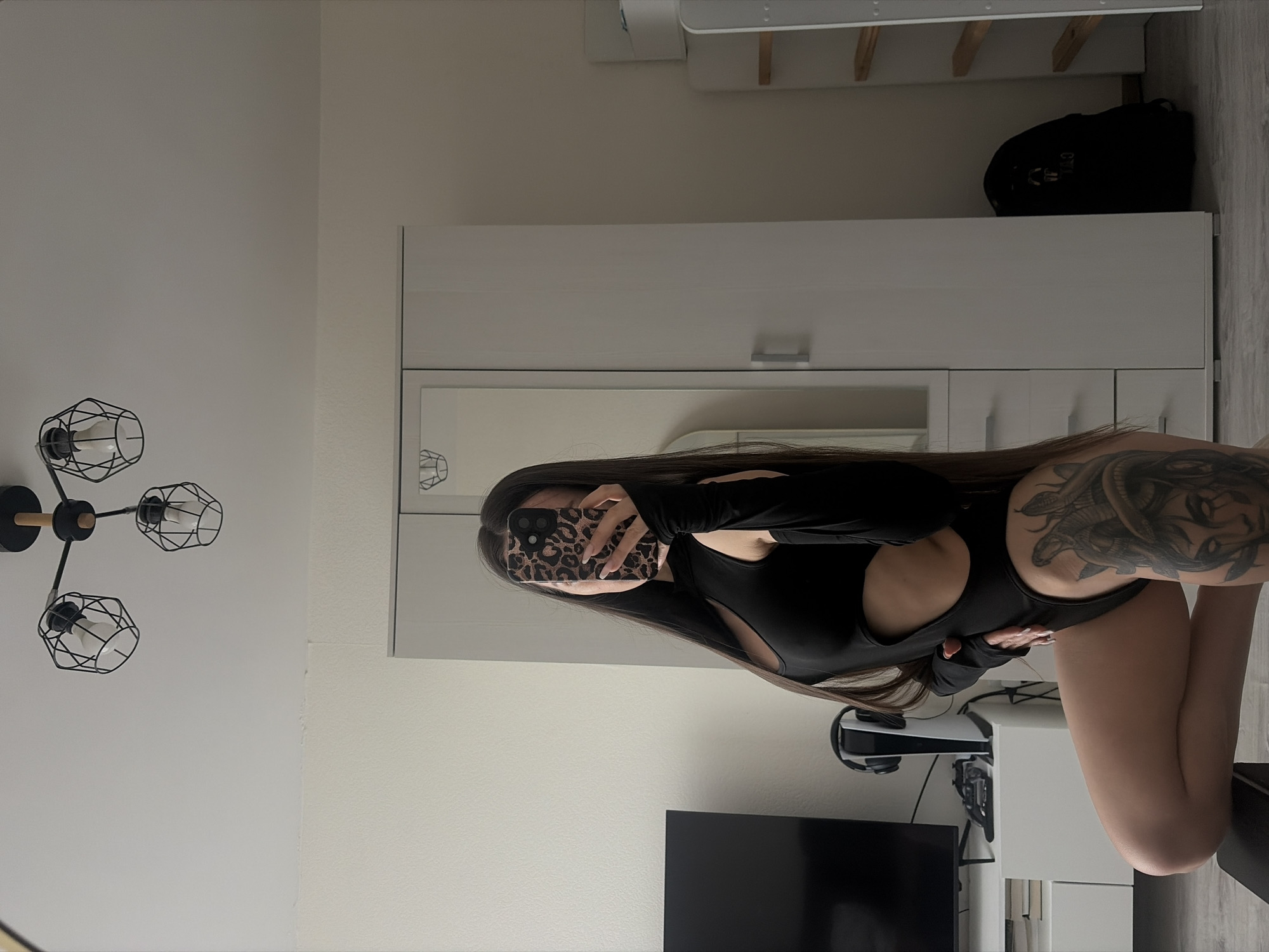 A woman is standing in front of a mirror, taking a selfie with her phone. She is wearing a black, form-fitting garment with cutouts. Her hair is long and brown. A large tattoo is visible on her arm and shoulder. The room appears to be a bedroom or living area, with a wardrobe, a television, a PlayStation console, and a modern light fixture.