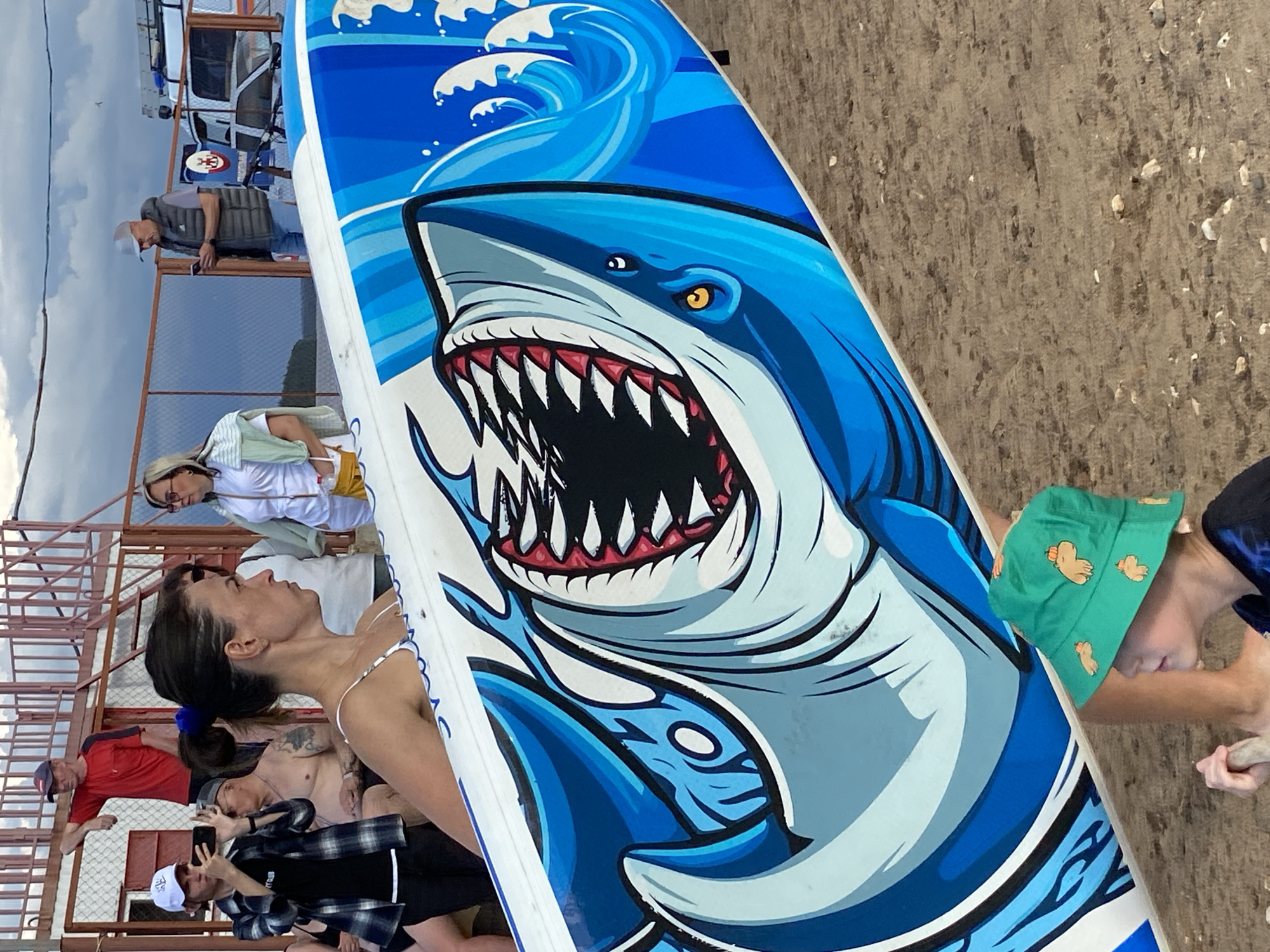 In Chaikovsky, Russia, a group of people are gathered near a beach or waterfront. The central focus is a surfboard with a graphic of a shark with its mouth open and teeth bared, surrounded by blue waves. The surfboard is positioned diagonally across the frame, obscuring some of the people.
Several individuals are visible, with varying degrees of focus and clarity. A woman in a white bikini is prominently featured in the foreground, looking away from the camera. To her left, a man in a red t-shirt and a baseball cap is also looking away. Another person, partially obscured, is seen with tattoos. Further back, a man in a grey vest and a white cap is looking at his phone. A woman in a white shirt and sunglasses is standing near a railing, holding what appears to be a bottle. On the right side of the frame, a person wearing a green bucket hat with cartoon pig faces is partially visible.
The setting appears to be outdoors, possibly a recreational area by the water, with a sandy or gravelly ground. There are metal railings and structures in the background, suggesting a pier or a viewing platform. The sky is overcast with clouds. The overall mood seems casual and relaxed, with people perhaps preparing for or enjoying an activity related to the surfboard. No discernible text is visible on the surfboard or clothing. The time of day appears to be daytime, given the lighting conditions.