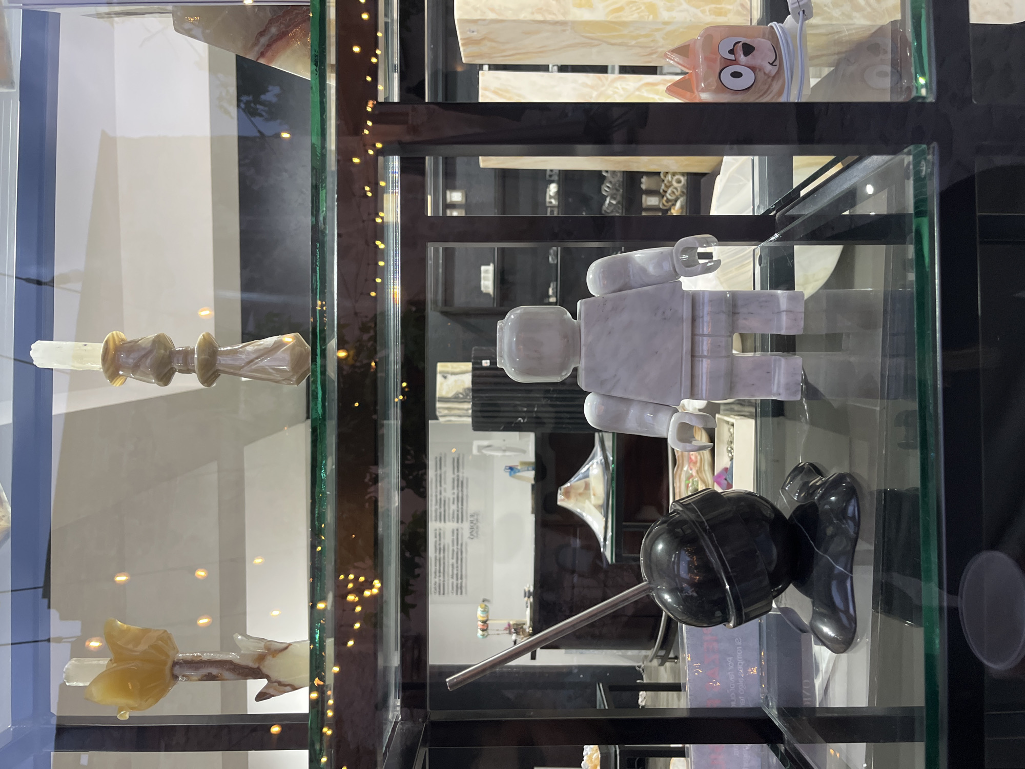 The image shows a display case in what appears to be a shop or gallery in Domingo Arenas, Mexico. The case is filled with various objects, likely artworks or decorative items. On the top shelf, there is a pink object with cartoonish features. The middle shelf holds a large white marble sculpture resembling a Lego figure. The bottom shelf contains a black sphere attached to a metal rod, possibly a sculpture, accompanied by a black object resembling a shoe. The overall lighting is bright, and the objects are displayed in a clear, organized manner.