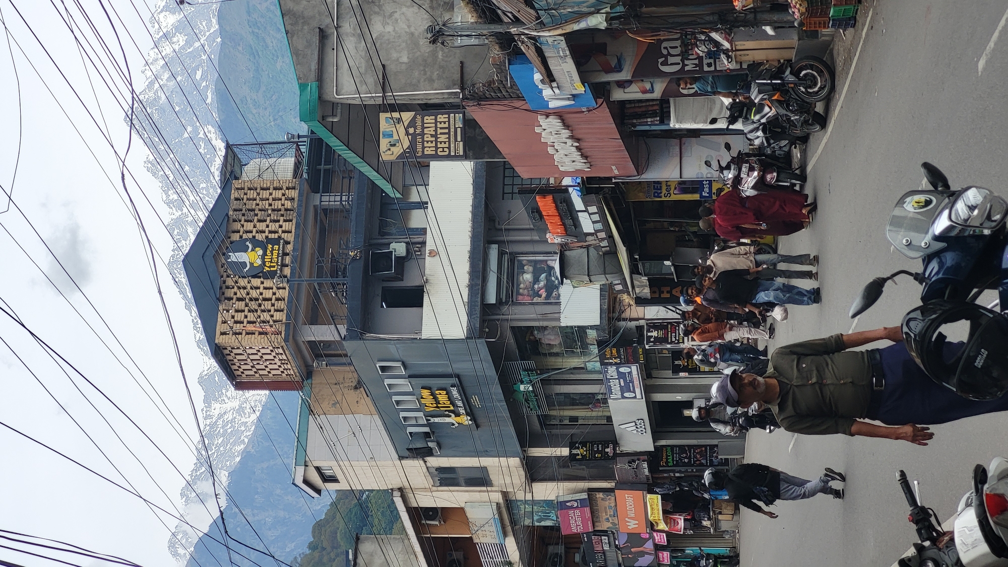 The scene is in the city of Dharamshala, India. In the foreground, two men are walking on the road. A motorcycle is parked in front of them. In the background, there is a building with many signs, including "Yellow Llama CAFE AND LOUNGE," "Gaurav Mobile REPAIR CENTER," and "AMERICAN TOURISTER." The Himalayan mountains are visible in the background.