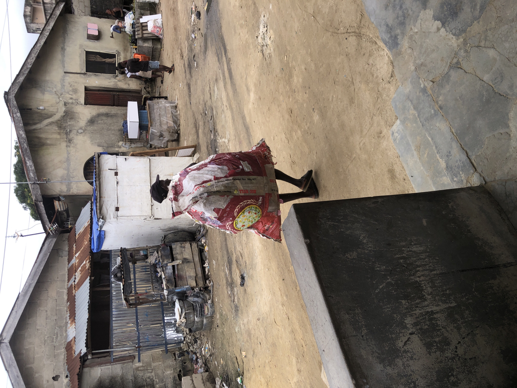 A person is walking away from the camera, shrouded in a large, red and white patterned bag. The bag, which appears to be made of woven material, covers the entirety of their body, obscuring their clothing and features. They are wearing dark sandals on their feet, and their legs are visible below the bag. The person is walking on a dirt path, with a weathered, grey concrete structure in the foreground. In the background, there are several buildings, some with corrugated metal roofs. One building in the middle ground is painted white and appears to be a temporary structure or stall. There are piles of debris and trash scattered around the environment, suggesting an area that is not well-maintained. Other people are visible in the background, some standing and talking near a building, and one person is sitting in a cart. The overall scene depicts a somewhat dilapidated urban environment, possibly in Ado, Nigeria, characterized by the presence of makeshift structures, scattered refuse, and everyday life unfolding in what appears to be a working-class or informal settlement.
