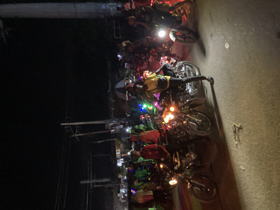 The image displays a crowded night street scene in Ura Nta Umuarandu, Nigeria. A large gathering of individuals, primarily male, are present, many positioned on or adjacent to numerous motorcycles. In the immediate foreground, a male subject wearing a yellow t-shirt, dark trousers, and dark sandals stands to the right of a dark motorcycle with its headlight illuminated; he holds a dark object, likely a mobile phone, to his right ear and wears a dark shoulder bag. To his left, another male subject in a red shirt and dark pants is seated on a similar dark motorcycle, also with an illuminated headlight.
Additional motorcycles, some with active headlights and others adorned with multi-colored LED string lights (blue, green, red), are visible throughout the frame. Further back, numerous people and motorcycles create a dense crowd. In the mid-ground, a person holds an open umbrella featuring distinct red and light-colored stripes. The background reveals blurred structures indicative of buildings or stalls lining the street, with multiple utility poles and overhead power lines silhouetted against the dark night sky. The ground surface is paved, possibly asphalt or packed earth, with some indeterminate debris in the closest foreground. Illumination is provided by vehicle headlights, decorative lights, and distant artificial sources, casting varied shadows across the scene.