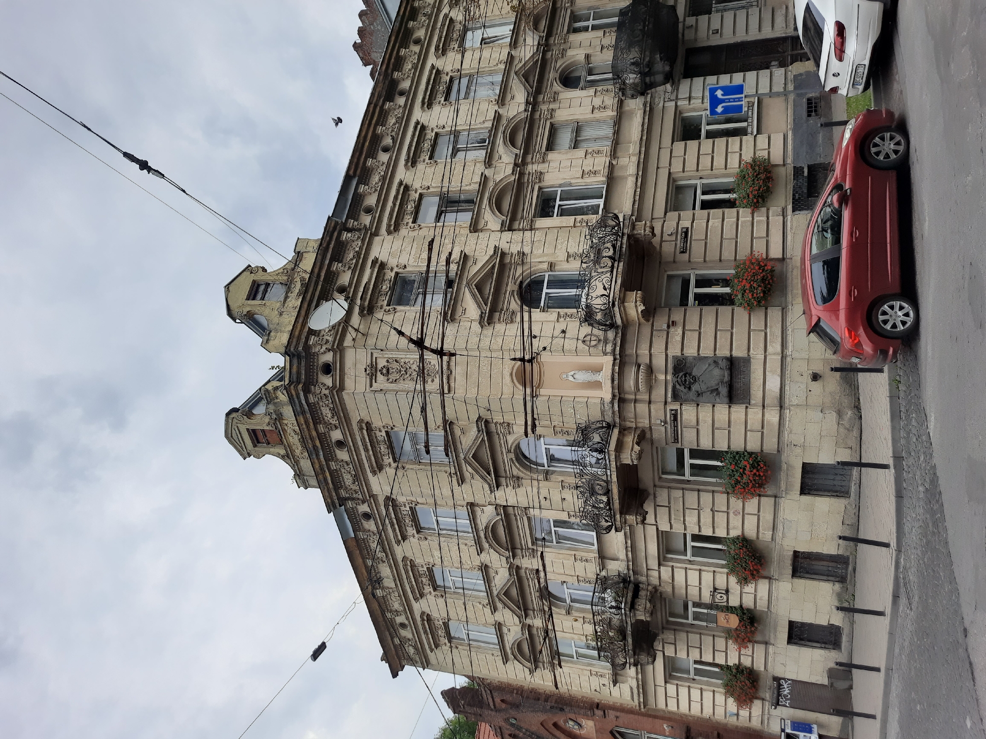 The image shows a corner of an ornate, multi-story building in Lviv, Ukraine, likely a residential or commercial building. The building's facade is made of light-colored stone with intricate architectural details, including cornices, decorative reliefs, and arched windows. Several wrought-iron balconies adorn the upper floors. There are also statues and plaques mounted on the building's exterior.
In front of the building, there is a sidewalk and a street. Two cars are parked on the street: a red hatchback and a white sedan. The sky is overcast, suggesting a cloudy day. The scene appears to be a typical urban streetscape. The building's architecture is indicative of late 19th or early 20th-century European styles. The presence of overhead wires suggests a developed urban area.