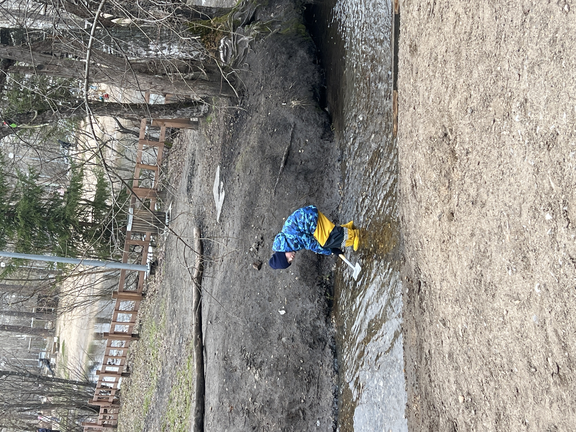 A young child is depicted playing in a shallow stream in an outdoor, likely park, setting in Perm, Russia. The child wears a blue and yellow patterned jacket, dark pants, bright yellow rubber boots, and a dark beanie hat. Bent over, they are holding a small white shovel, seemingly digging or scooping something from the water or the muddy bank.
The immediate surroundings feature a dark, muddy bank adjacent to the stream on one side, and a lighter, sandy or gravelly bank on the other. Bare, branch-heavy deciduous trees dominate the background, with glimpses of evergreen trees and a wooden fence running along the far bank. A path or open area is visible further in the background.
The absence of leaves on the trees, the muddy ground, and the child's warm attire (jacket, hat, and rubber boots) suggest a cool or cold season, such as late autumn, winter, or early spring. The lighting is soft and even, indicating an overcast day. The child appears focused and engrossed in their activity. No text is visible in the image.