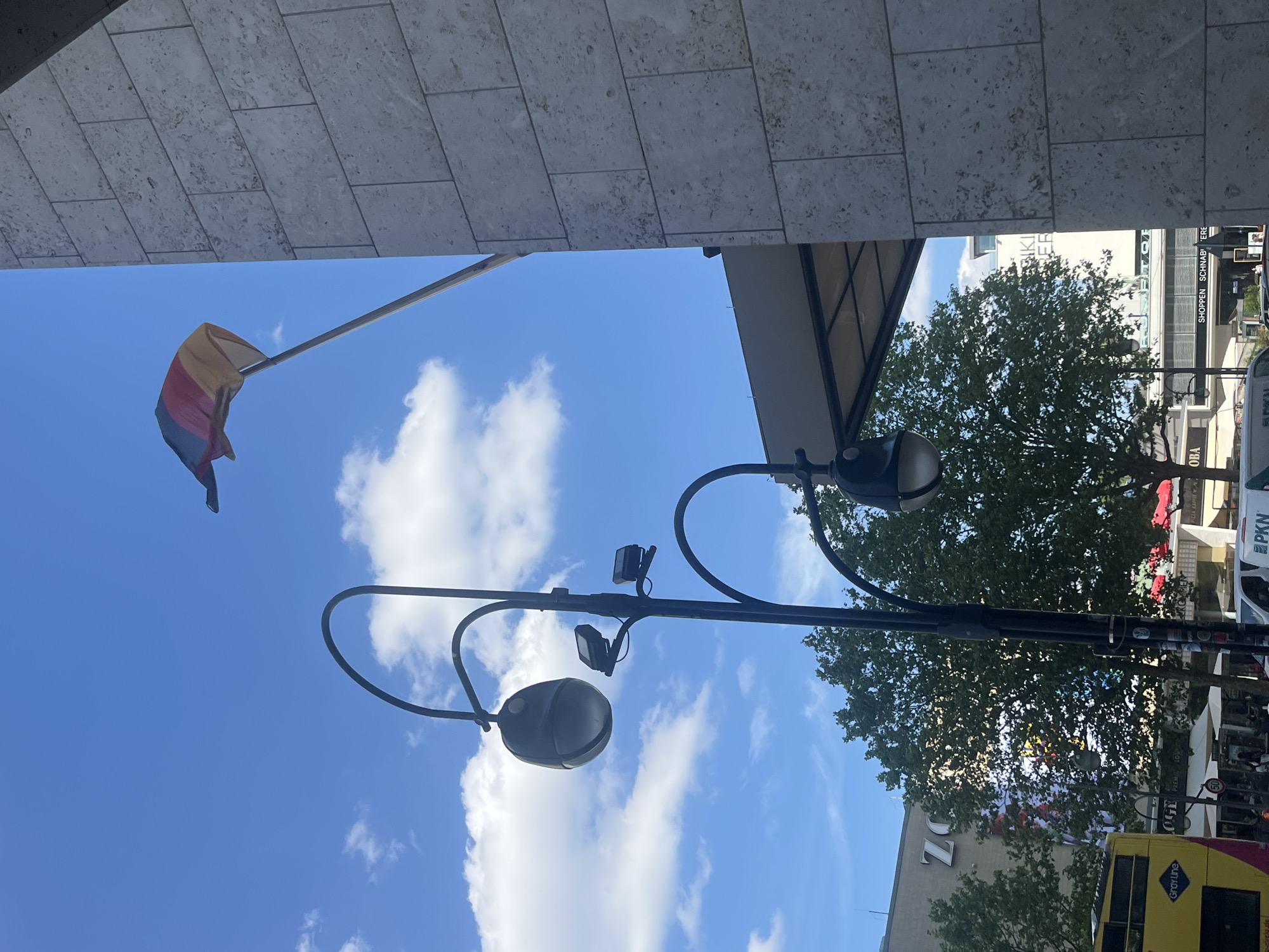 This is a vertically oriented, outdoor photograph taken from a low angle looking up at a bright blue sky with scattered white clouds. A street lamp with multiple lights and cameras is prominently featured in the foreground, its dark metal structure arching across the frame. A flag, with horizontal stripes of yellow, pink, and blue, is visible flying from a pole on the left side of the image.
In the background, a building with large windows and beige stone cladding is partially visible at the top. Further in the distance, lush green trees are in full leaf, suggesting a warm season, possibly summer. Below the trees, a street scene unfolds with glimpses of buildings, shops, and a yellow double-decker bus. Some text is visible on the buildings, including "ZO", "Gray Line", "LOGE", "PKN", "OBA", and "SHOPPEN SCHNAB' 'ERE". The overall impression is a sunny day in a city, likely Berlin, Germany, with a festive or pride-related element suggested by the flag. The low angle of the shot emphasizes the sky and the architectural elements above.