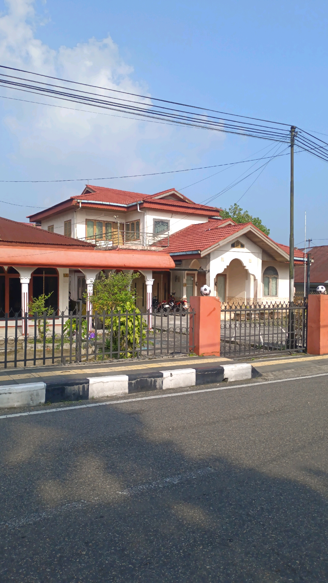 The image depicts a large, two-story house with a red tile roof, characteristic of Indonesian architecture. The house features a prominent white facade with arched windows and decorative pillars supporting a covered walkway. A dark metal fence encloses the property, separating it from the paved street in the foreground. Several motorcycles are parked under the covered walkway, suggesting a residential setting. On top of two orange pillars flanking the gate, there are decorative soccer balls. The sky is clear blue with a few scattered clouds. The scene is captured from a street-level perspective, emphasizing the house and its immediate surroundings. The image appears to be taken in Padang Panjang, Indonesia, as indicated by the architectural style and the general environment, which aligns with typical urban or suburban landscapes in that region.