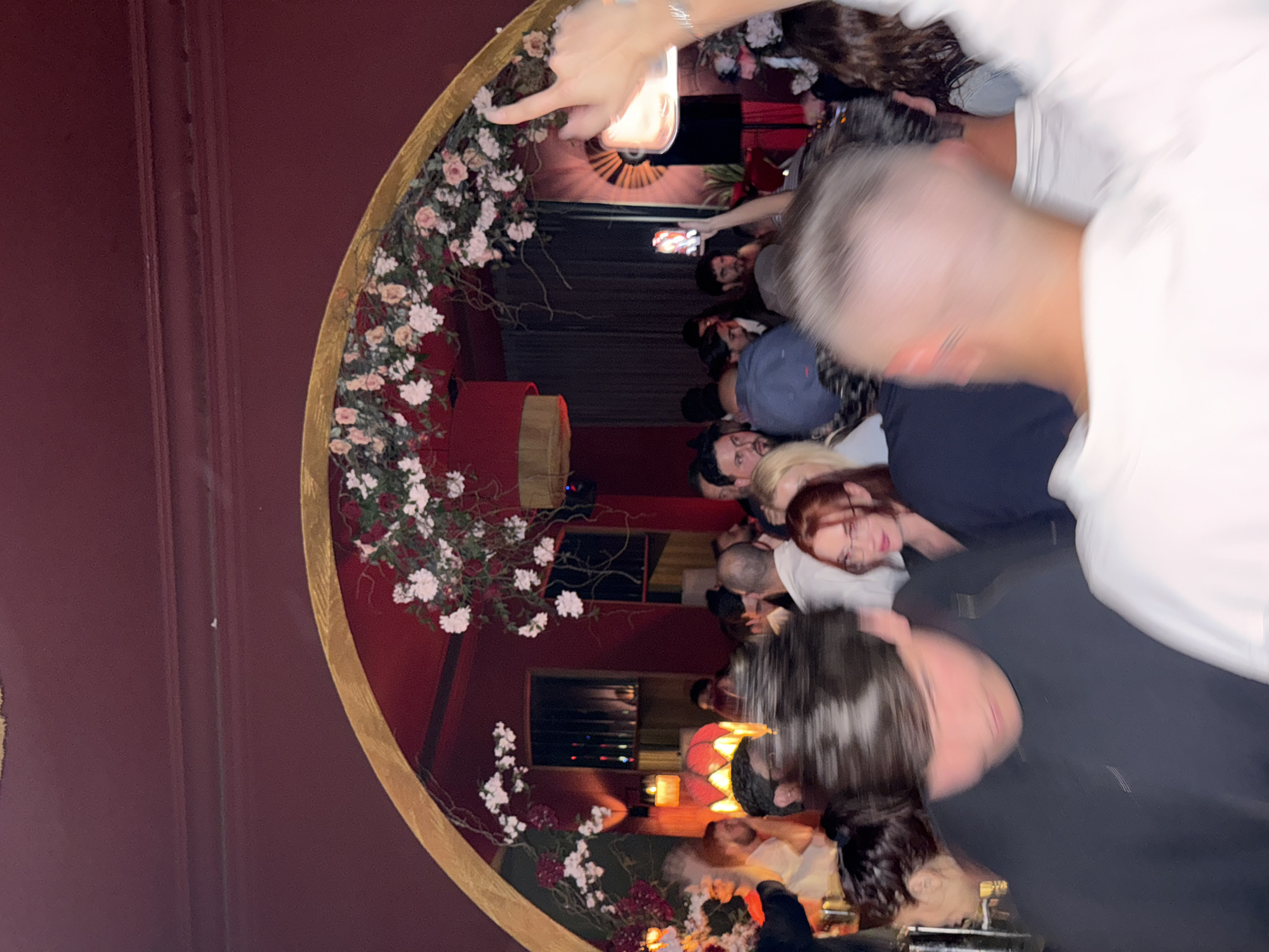 The scene captured in the image appears to be a crowded gathering inside a venue in Yerevan, Armenia. The venue has a distinctive decorative element: a large, ornate, circular structure adorned with floral arrangements that resembles a stylized mirror or portal. The backdrop is a dark red wall.
Several people are visible, engaging in what seems like social interaction. Many appear to be looking at or interacting with mobile phones, possibly taking pictures or videos. The lighting is warm and suggests a lively atmosphere. The overall impression is one of a social event or party.