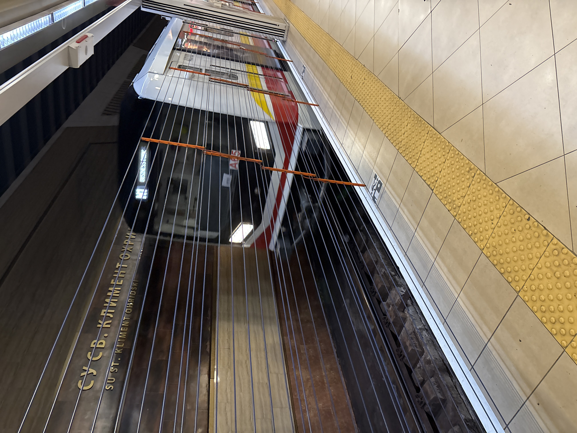 The image displays a modern metro station platform and tracks from an elevated, angled perspective. A metro train, partially visible on the right, is stationary alongside the platform. The train features a white body with prominent red and yellow horizontal stripes.
The platform is tiled with light-colored square tiles, and a bright yellow tactile paving strip, featuring raised dots, runs diagonally across it, marking the platform edge. A safety barrier, constructed from numerous thin, vertical metal wires and several orange horizontal support bars, separates the platform from the train tracks.
On a dark, highly reflective surface to the left, which might be a display panel or a polished wall, the name of the station is visible in two languages. In Cyrillic, it reads "СУ СВ. КЛИМЕНТ ОХРИДСКИ", and in Latin script, "SU ST. KLIMENT OHRIDSKI". These gold-colored letters are reflected on the glossy surface. Further left, a small white box with a red button is mounted on a lighter-colored wall.
Beyond the safety barrier, the train tracks are visible, laid on a bed of dark ballast. The scene is illuminated by artificial lighting, typical of an underground metro environment. No individuals are present, focusing the view entirely on the station infrastructure. The visible text confirms the location as the "SU St. Kliment Ohridski" metro station in Sofia, Bulgaria.