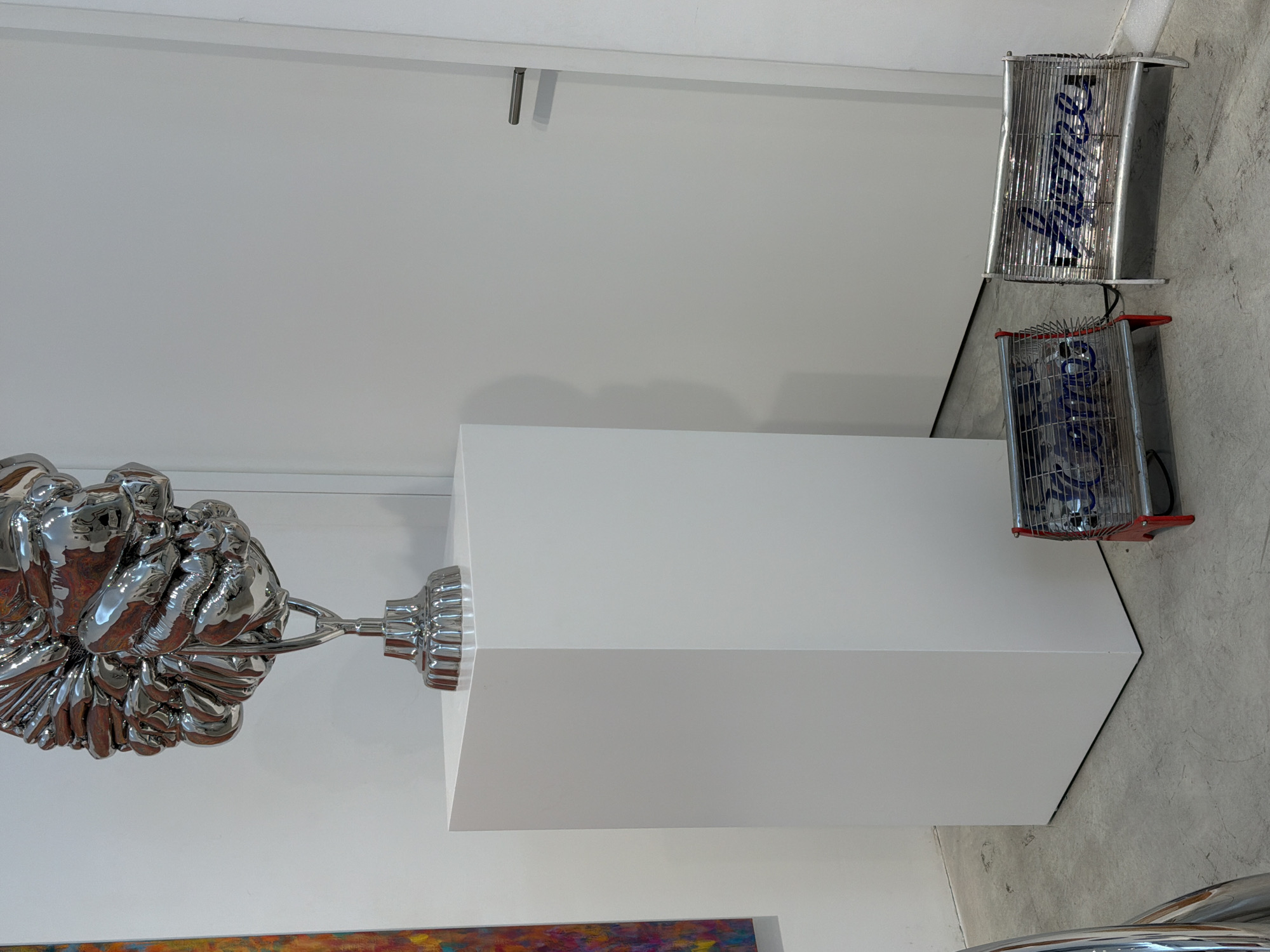 In a minimalist gallery space, a polished chrome sculpture with intricate, bulbous forms hangs suspended from the ceiling, its reflective surface catching the light. The sculpture is attached to a slender rod that connects to a circular, fluted base. To the right of the sculpture, a white pedestal stands, its sharp geometric lines contrasting with the organic curves of the artwork.
Above the pedestal and to the right, two vintage-style electric fans are mounted on the wall. The fans are housed in chrome cages with red accents and feature blue neon tubing that spells out text. The upper fan displays the word "JEWISH" in blue, while the lower fan shows "MEN." The fans are positioned vertically against a textured concrete wall, suggesting an installation piece that incorporates industrial elements into an artistic display. The overall scene is brightly lit, typical of an indoor exhibition space. The location is Tel-Aviv, Israel.