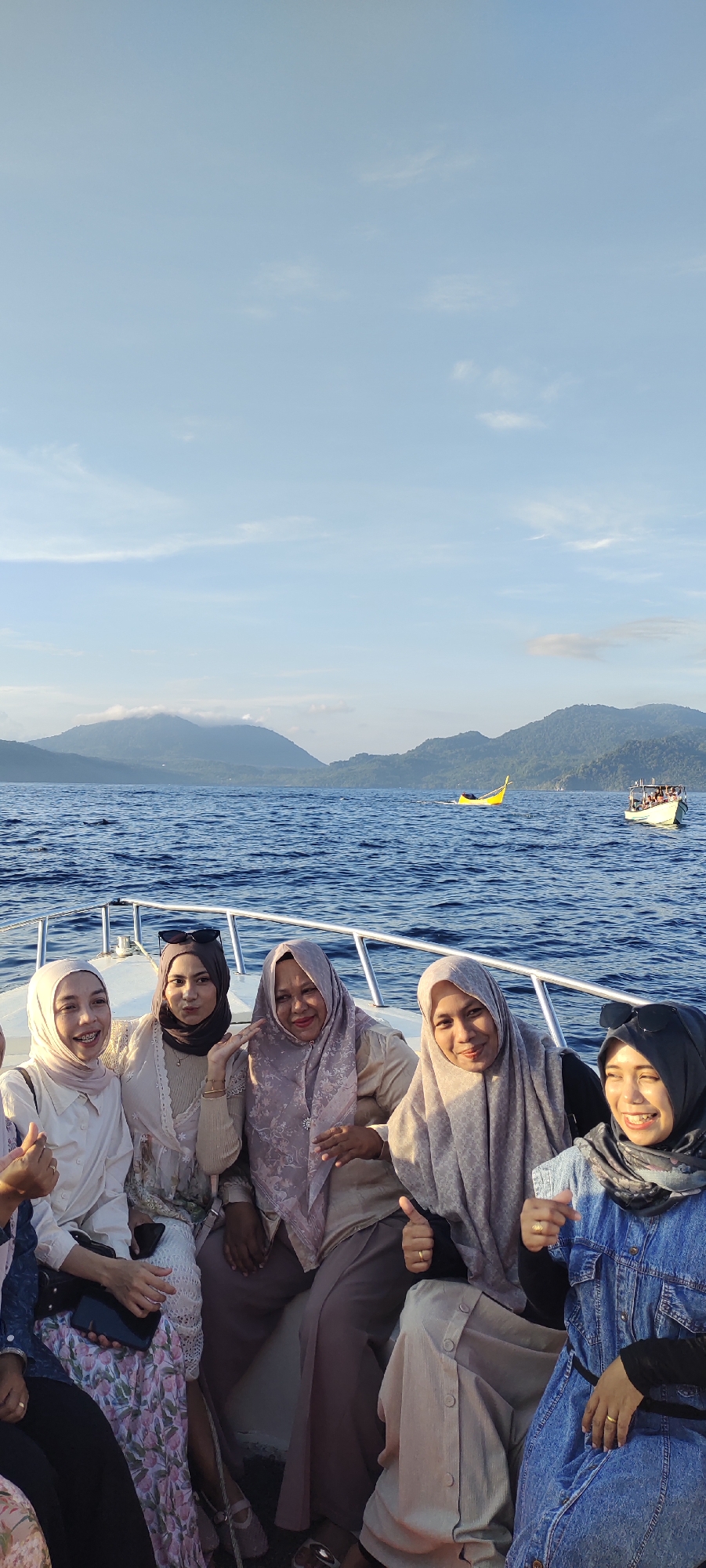 Six women are seated on the deck of a boat, posing for a photo. The boat is on the water with a backdrop of lush green hills and a blue sky with scattered clouds. In the distance, two other boats are visible, one yellow and one white. The woman on the far left wears a cream-colored hijab and shirt. Next to her, a woman with a dark brown hijab and a light beige top smiles and gives a thumbs-up. The third woman from the left, wearing a light purple hijab and a beige top, also smiles and gestures with her hand. The fourth woman from the left, in a light beige hijab and top, smiles and gives a thumbs-up. The fifth woman, wearing a dark grey hijab and a denim jacket, smiles and raises her hand. The sixth woman, partially visible on the far right, is wearing a blue denim outfit and a dark hijab, and appears to be smiling. The scene is likely taking place in the waters off the coast of Sabang, Indonesia, given the tropical environment and the presence of traditional Indonesian boats.