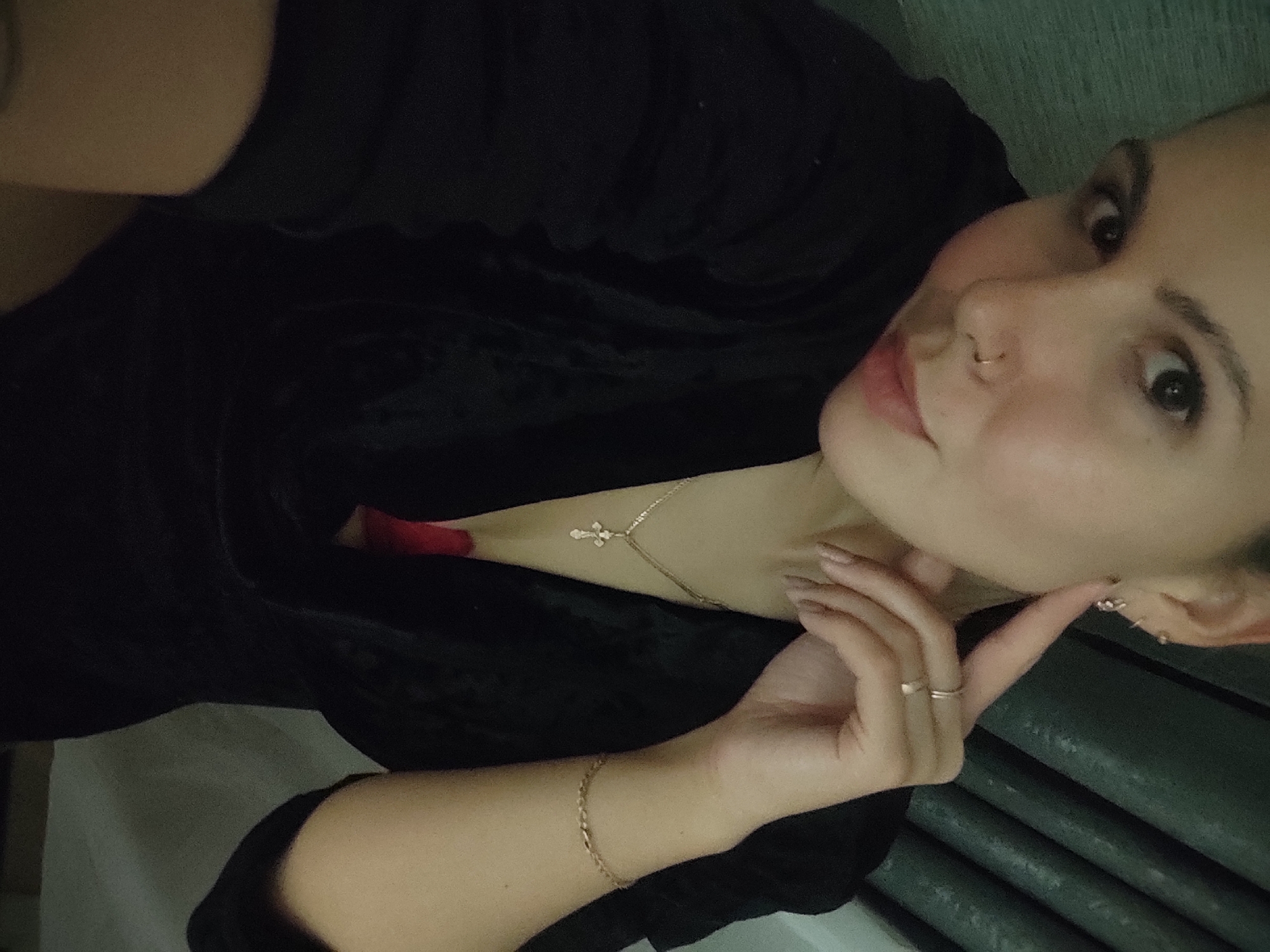 A fair-skinned woman with light brown hair pulled back is angled in a selfie-style shot, gazing at the camera. She has a nose ring in her left nostril and multiple earrings in her left ear. She wears a black velvet top with a hint of red fabric visible beneath. A gold necklace with a cross pendant adorns her chest. She has a bracelet on her left wrist and rings on her left hand, which is positioned near her face. The background includes what appears to be white fabric and a vertically ribbed surface, potentially a wall or door. The location is likely indoors, but the image provides no specific information to confirm its location in Chaikovsky, Russia.