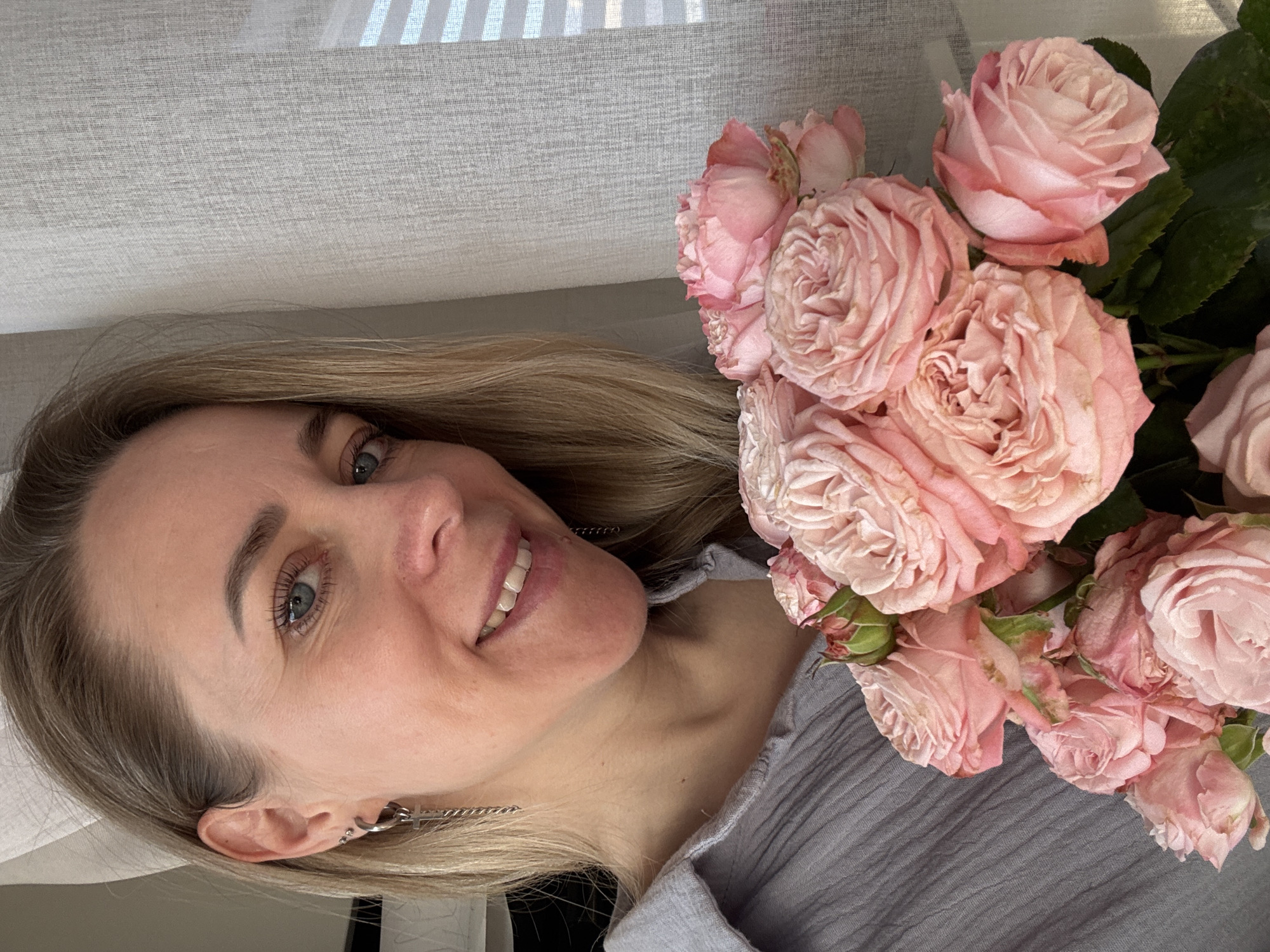 A fair-skinned woman with blonde hair and blue eyes smiles at the camera, holding a bouquet of pink roses. She has multiple earrings in her left ear. She wears a grey top. A light grey curtain is visible in the background. The city of Chaikovsky, Russia is not discernible from the provided image.
