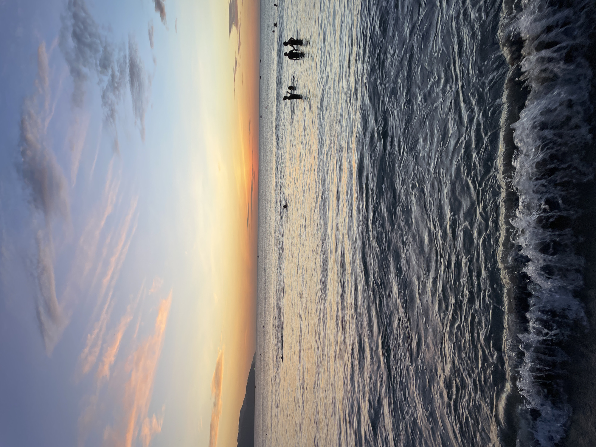 This image captures a sunset scene at My Khe Beach in Đà Nẵng, Vietnam. The sky is a gradient of colors, transitioning from pale blue at the top to shades of pink and orange near the horizon, with wispy clouds scattered throughout. The ocean surface reflects the sky's colors, creating a shimmering effect. In the foreground, small waves are breaking on the shore, with white foam. Several silhouetted figures, appearing to be people, are visible in the water, some wading and others possibly swimming. A distant shoreline with hills is visible on the left side of the frame. The overall impression is one of a serene evening at the beach.