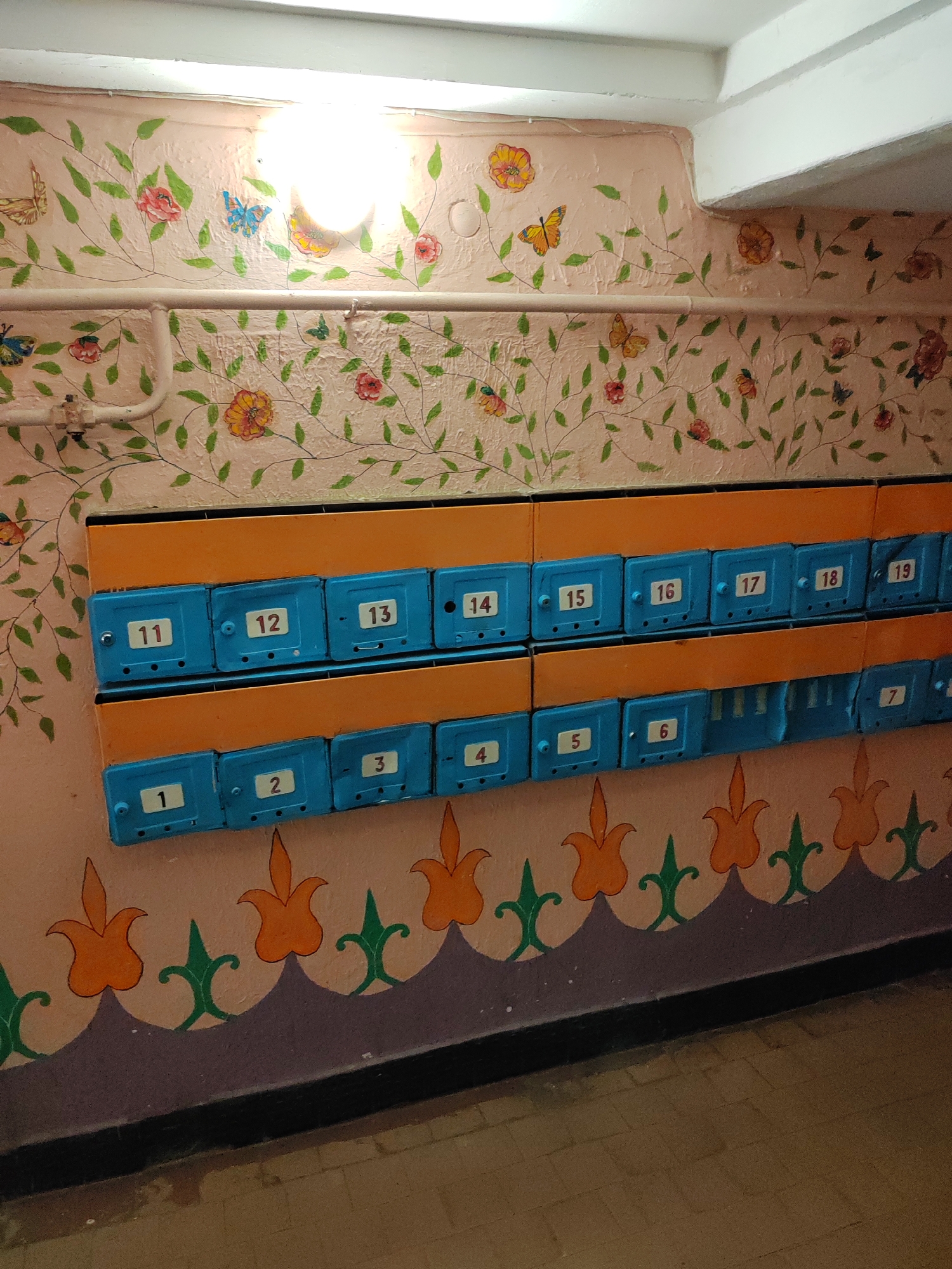 The image shows a hallway with a floral and butterfly motif painted on the walls. There are two rows of blue mailboxes mounted on an orange panel. The mailboxes are numbered sequentially from 1 to 19, with some numbers missing. The lower row of mailboxes is partially obscured by a decorative border painted on the wall. The floor is tiled with brown bricks. The lighting appears to be from an overhead fluorescent fixture. This scene is consistent with the entrance of a residential building in Brest, Belarus.