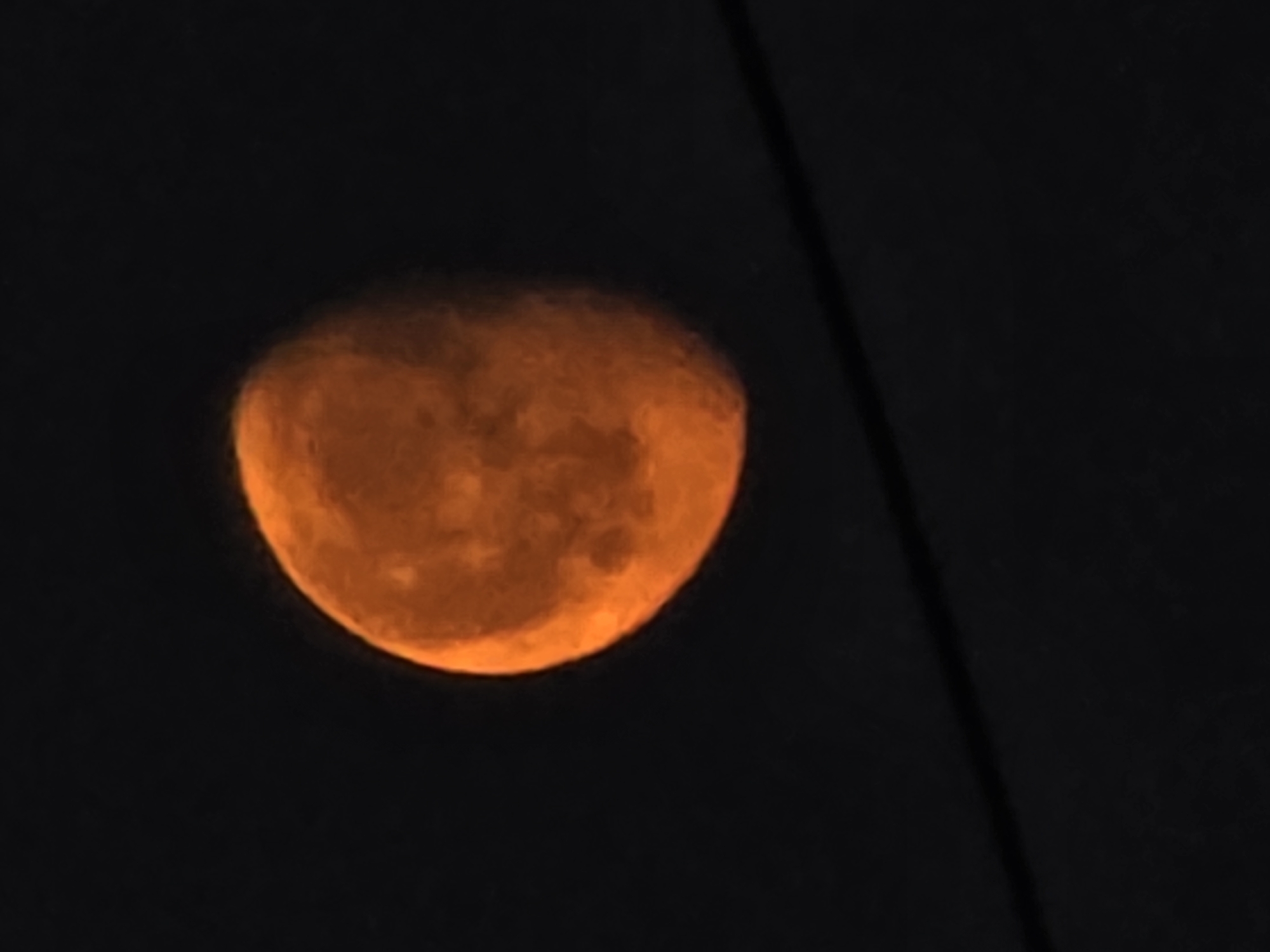 The image depicts an orange moon against a dark background. A diagonal dark line crosses the right side of the image. The location is unknown. There are no people visible.