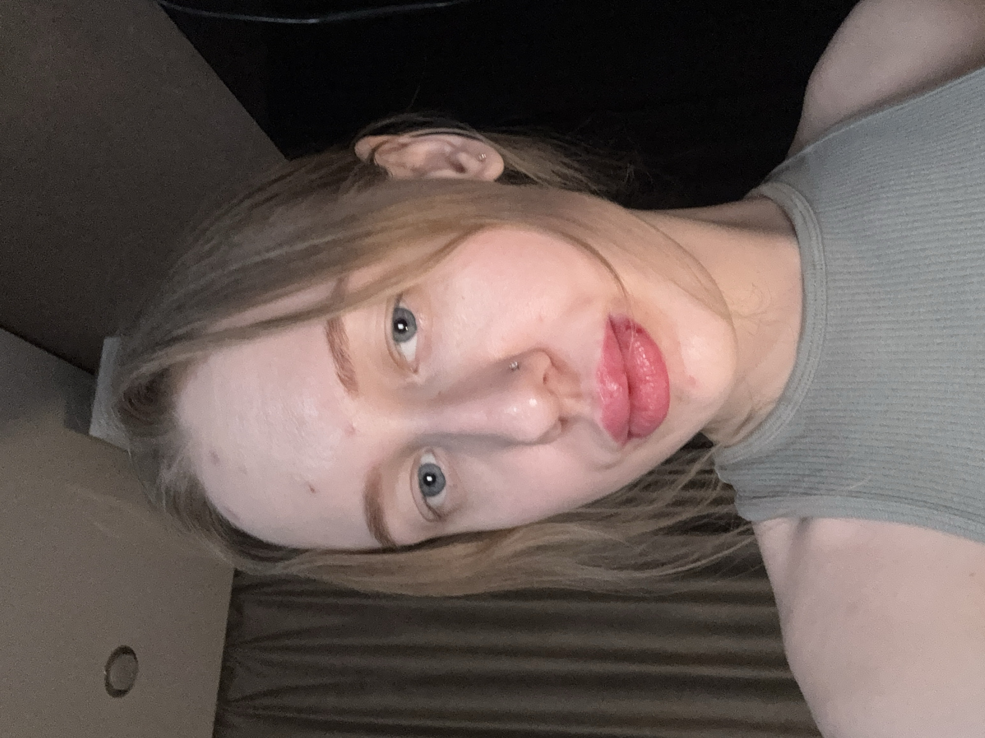 A light-skinned woman with light brown hair and blue eyes is shown in a close-up self-portrait. She is wearing a light green tank top. She has a pierced nose and a pierced ear. The background includes a dark area, and a light-colored, possibly wooden or fabric-covered surface. The scene does not provide enough information to indicate the location in Rostov-on-Don, Russia.