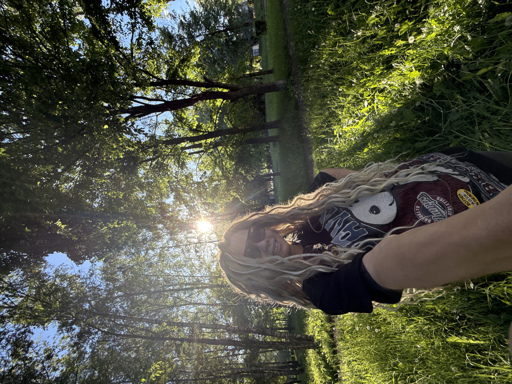 The image is a vertical full-frame selfie taken outdoors in a park or green space within the city of Rusanovo, Russia. The central subject is a person, appearing to be female, smiling broadly and looking directly at the camera. She wears dark sunglasses, obscuring her eyes, and has long, prominent light blonde and multi-toned dreadlocks or braids that frame her face and extend over her chest and shoulders. She is wearing a dark short-sleeved t-shirt with a maroon section featuring a graphic of a black-and-white figure and text including "BROOKLYN CHAMPS ATHLETICS" and a yellow patch with "ROTHER B.O.T.R ON THE ROAD". Her right arm is visible in the lower right foreground, extending towards the camera.
The environment is lush and green, characteristic of a sunny day. Strong sunlight originates from the upper right background, creating a distinct sunburst and lens flare, backlighting the subject and brightly illuminating the surrounding foliage. The foreground is covered in tall, green grass, interspersed with white dandelion seed heads.
The background features a dense canopy of deciduous trees with vibrant green leaves, through which patches of clear blue sky are visible. Some trees exhibit lighter bark consistent with birch species on the left side, while others have darker trunks. In the far background, particularly on the right, less dense foliage suggests a more open area or a path, potentially with faint indications of distant structures.