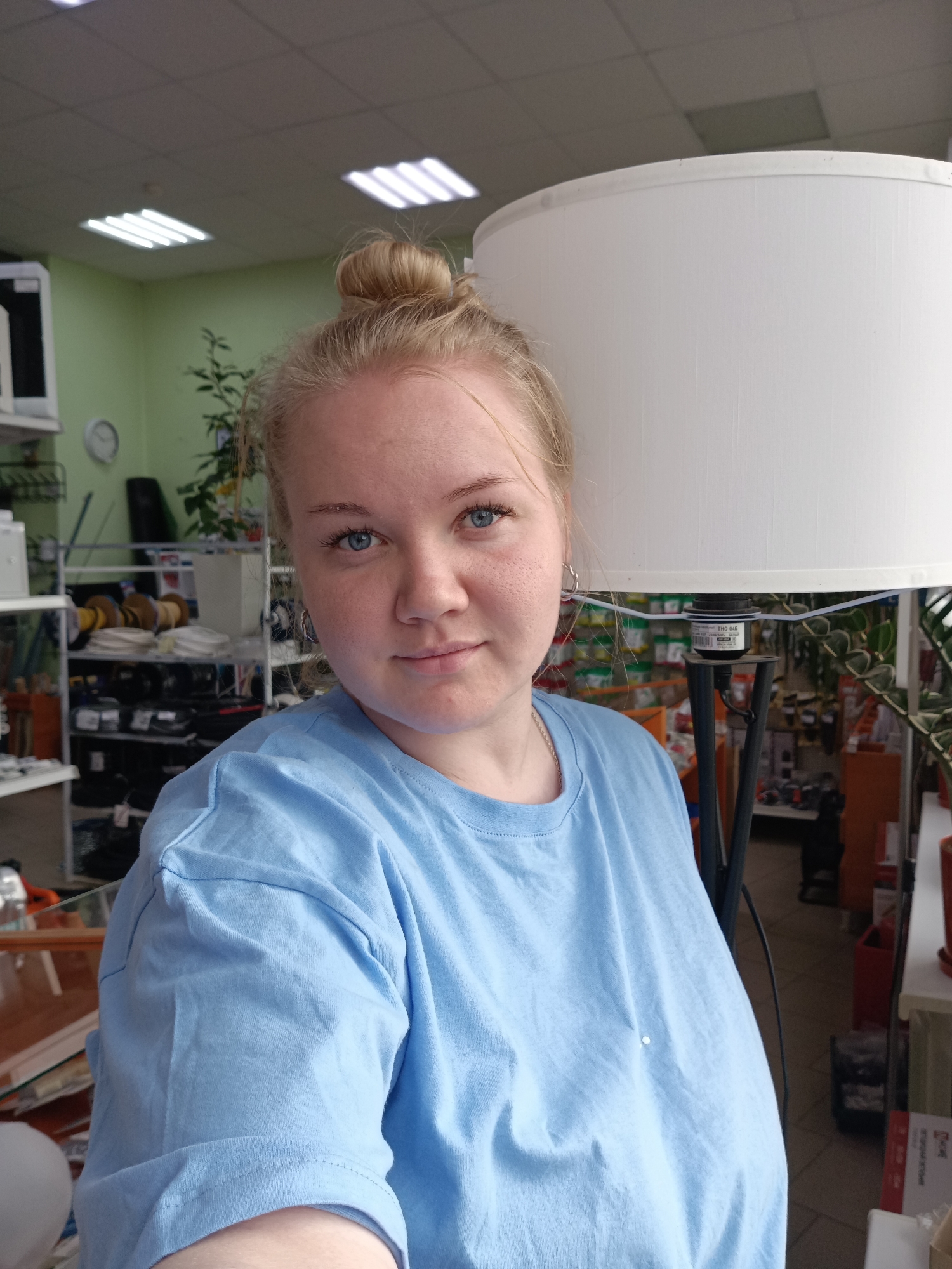 A young woman with fair skin, blue eyes, and blonde hair styled in a bun is the primary subject. She is wearing a light blue T-shirt and silver hoop earrings. She is indoors, likely in a retail environment, as suggested by the shelves in the background filled with various merchandise like spools of wire and other hardware items. A floor lamp with a large white lampshade is partially visible to her right. The background also includes green walls, fluorescent lighting, and plants. The image appears to be taken in a shop, possibly a hardware or home goods store, in Фоки, Russia.