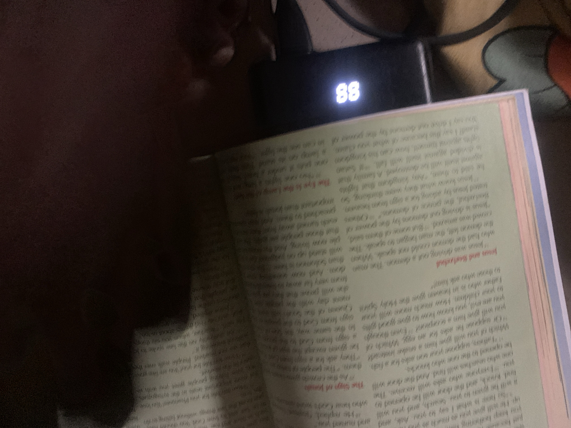 The image shows an open book with text and an electronic device with a digital display. The book is open to a page with a title in bold red font that reads "The Son of Moses." The text in the book is small and in English. The electronic device has a black casing and displays the number "88" in white light. The device is placed on a surface that appears to be a table. The background is dark and out of focus, suggesting a dimly lit environment. There is no identifiable information or context to suggest the location is Jalingo, Nigeria.