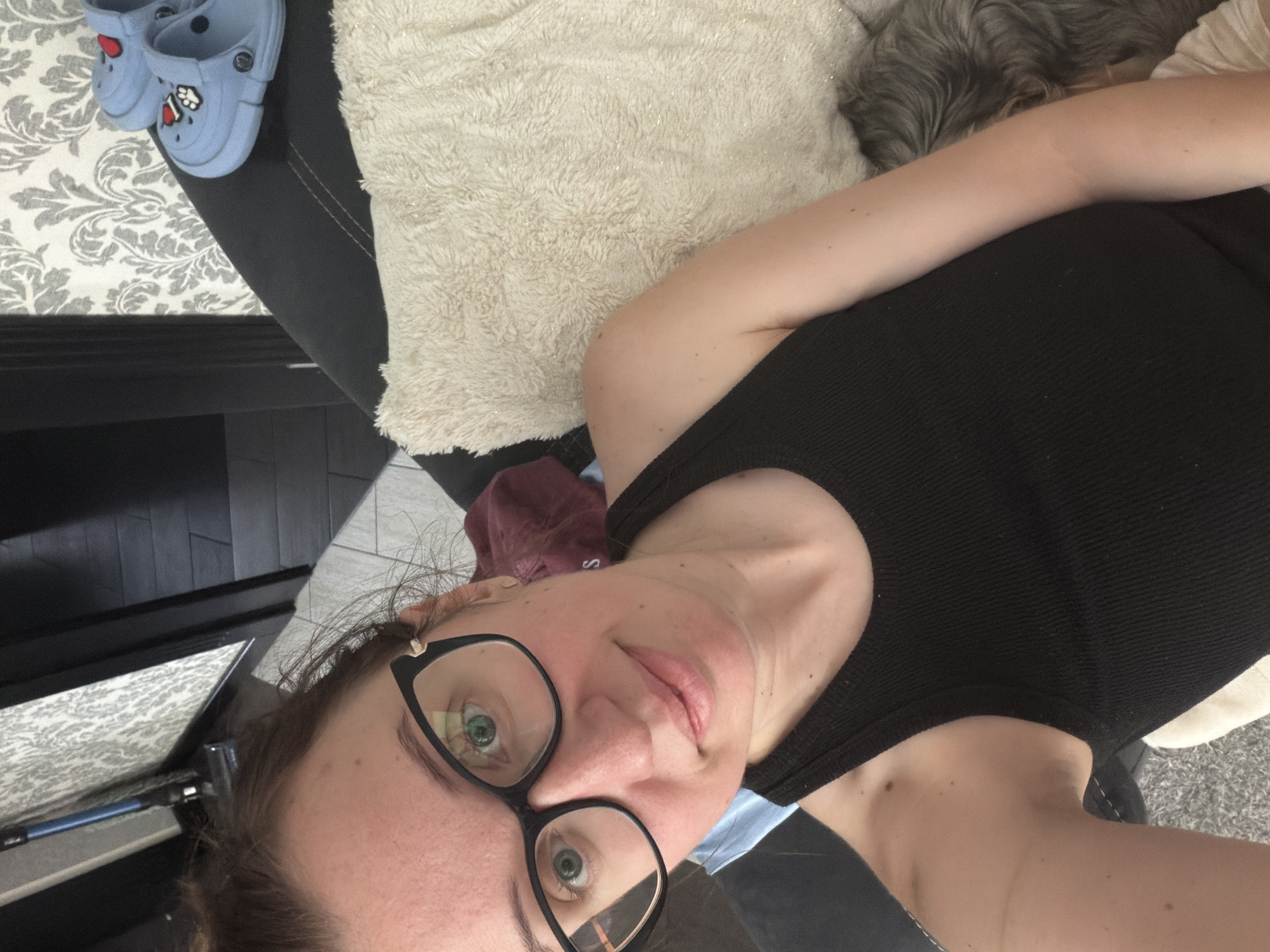 The image shows a close-up, high-angle, selfie-style shot of a woman lying on a black couch. She is wearing a black tank top and glasses. A fluffy cream-colored pillow is behind her to the right. Partially visible in the background are blue Crocs shoes with a red heart and paw print design, gray patterned wallpaper, and dark flooring. A vacuum cleaner is visible in the upper left corner. The woman's hair is pulled up in a bun. Her right arm is resting on the couch. The image does not contain any visual cues to suggest a specific location within the city of Bila Tserkva, Ukraine.