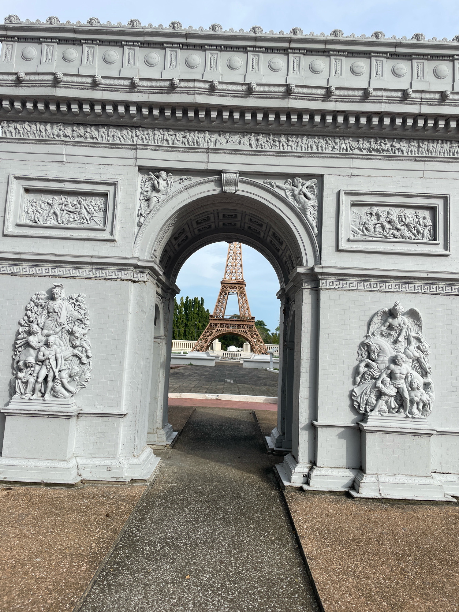 The image shows a miniature replica of the Arc de Triomphe in Paris, with a miniature Eiffel Tower visible through its arch. The replica is painted white and features ornate carvings on its facade, depicting figures and scenes. The Arc de Triomphe replica is situated within a park-like setting with paved walkways and some trees in the background. This scene is located in Pattaya City, Thailand, specifically at the "Mini Siam" attraction, which is a miniature replica park featuring famous landmarks from around the world. The sky is visible above, and the overall lighting suggests it is daytime, likely with moderate cloud cover. There are no people visible in the image.