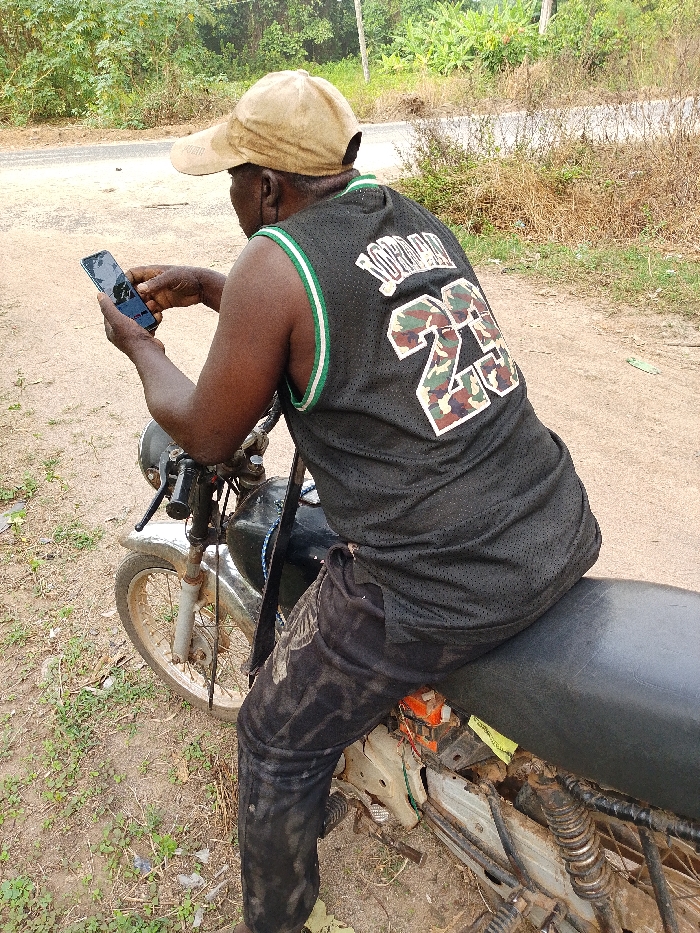 An African man wearing a tan baseball cap and a black Michael Jordan tank top sits on a motorcycle, looking at his phone. The motorcycle is old and shows signs of wear and tear. The man is wearing black pants. The background shows a dirt road with vegetation on either side. A paved road is visible in the distance.
