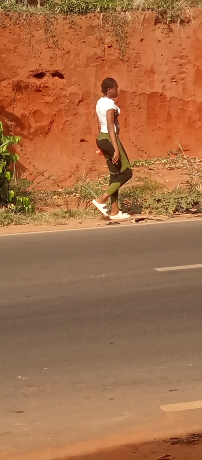 An outdoor scene in Ogidi, Nigeria, depicts a lone individual walking leftward along the edge of a paved road. The individual is a female, seen in profile, wearing a white short-sleeved shirt, olive green and black patterned leggings, and white flat sandals. Her hair is styled in dark braids. To her left, partially obscured, is green foliage. Behind her, extending across the upper two-thirds of the image, is a distinct reddish-brown earth embankment, uneven in texture, with sparse green and dry vegetation growing on its surface and at its base. The paved road surface occupies the lower third of the image, featuring a faint white broken line marking. The lighting suggests daylight conditions.