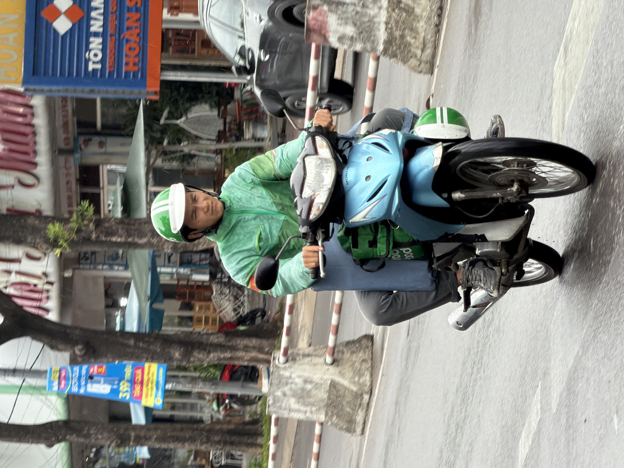 A person is riding a blue scooter on a wet city street in Nha Trang, Vietnam. The rider is wearing a green jacket, blue pants, and a green and white helmet. They are looking forward with a neutral expression. The scooter has a large green bag attached to the back. The street is lined with trees and buildings. There are concrete barriers and a striped pole in the foreground, indicating it might be a road under construction or a protected area. A dark-colored car is visible on the right side of the frame, also on the street. The sky is overcast, contributing to the wet appearance of the road. There are various signs and advertisements visible on the buildings in the background.