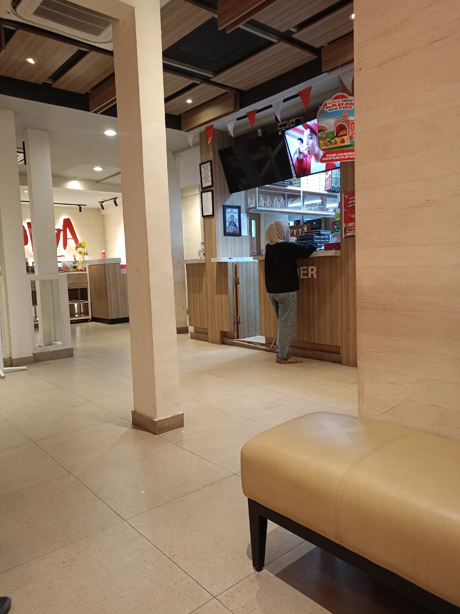 The image depicts the interior of a fast-food restaurant, likely a Pizza Hut, in Batu, Indonesia. In the foreground, a tan leather bench with dark wooden legs is visible on a tiled floor. In the mid-ground, a person wearing a light-colored hijab and grey jeans stands at a counter, facing away from the camera. They appear to be interacting with the counter, possibly ordering or making a payment. The counter is made of wood and has a sign with "ER" visible. Behind the counter, shelves with glasses and other items are present, along with a television screen displaying an advertisement for a pizza promotion, featuring a young person eating pizza and the text "MAKIN SERUI ADA PLAY-DOH UNTUK SI KECIL." To the left of the counter, large white pillars and part of the restaurant's branding, a red "A," are visible. The ceiling features wooden panels and recessed lighting. The overall environment suggests a casual dining setting.