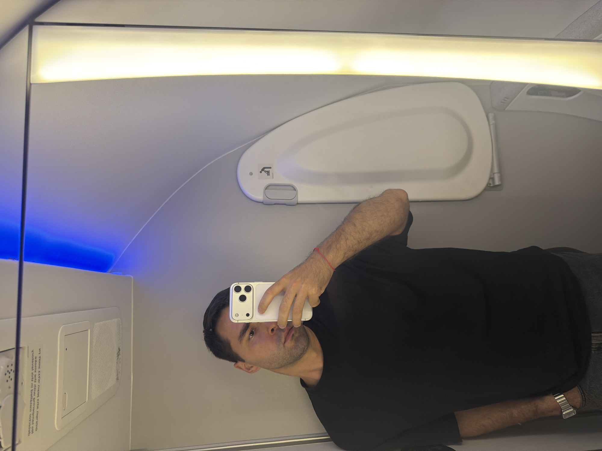 A man wearing a black t-shirt, grey pants, and a watch is taking a selfie with a white smartphone inside an airplane cabin. Overhead storage is visible with a white door and a light fixture. The environment appears to be an airplane cabin with overhead storage and interior lighting. No indications suggest this is in Tzafria, Israel.