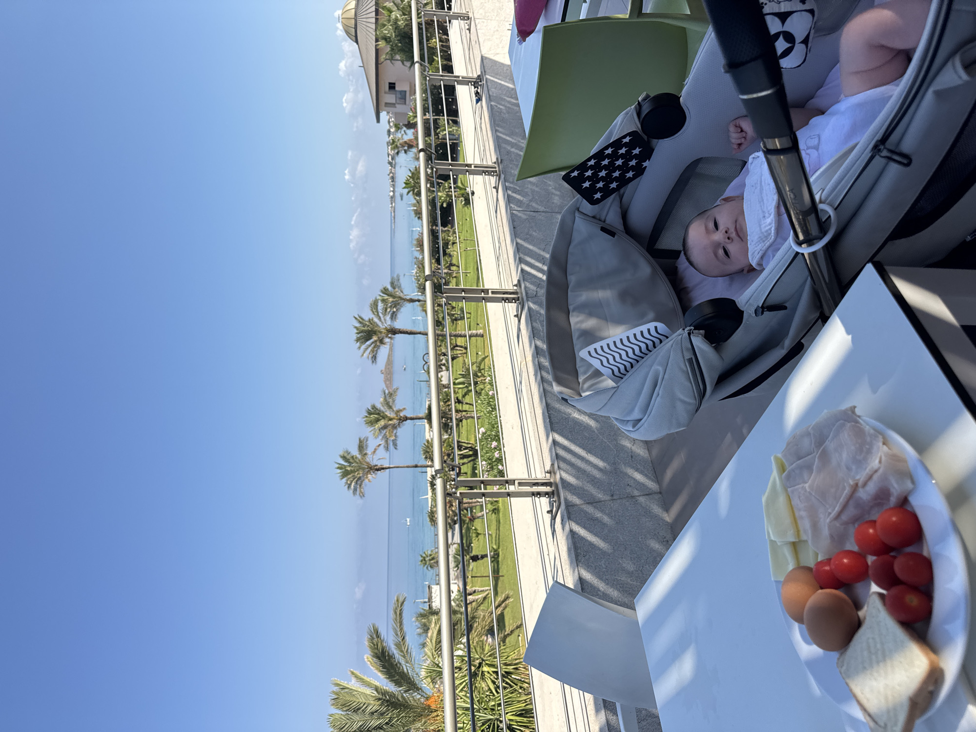 In Isola delle Femmine, Italy, a baby lies in a stroller on an outdoor terrace overlooking the Mediterranean Sea. The baby is wearing a white onesie and bib, with a black and white star-patterned accessory attached to the stroller. In the foreground, a plate of breakfast items sits on a white table, featuring two brown eggs, several cherry tomatoes, slices of cheese, ham, and a piece of toast. The terrace has white tables and chairs, a metal railing, and stone flooring. Beyond the terrace, palm trees and green grass are visible, leading to the turquoise sea with boats in the distance. The sky is a clear, bright blue.