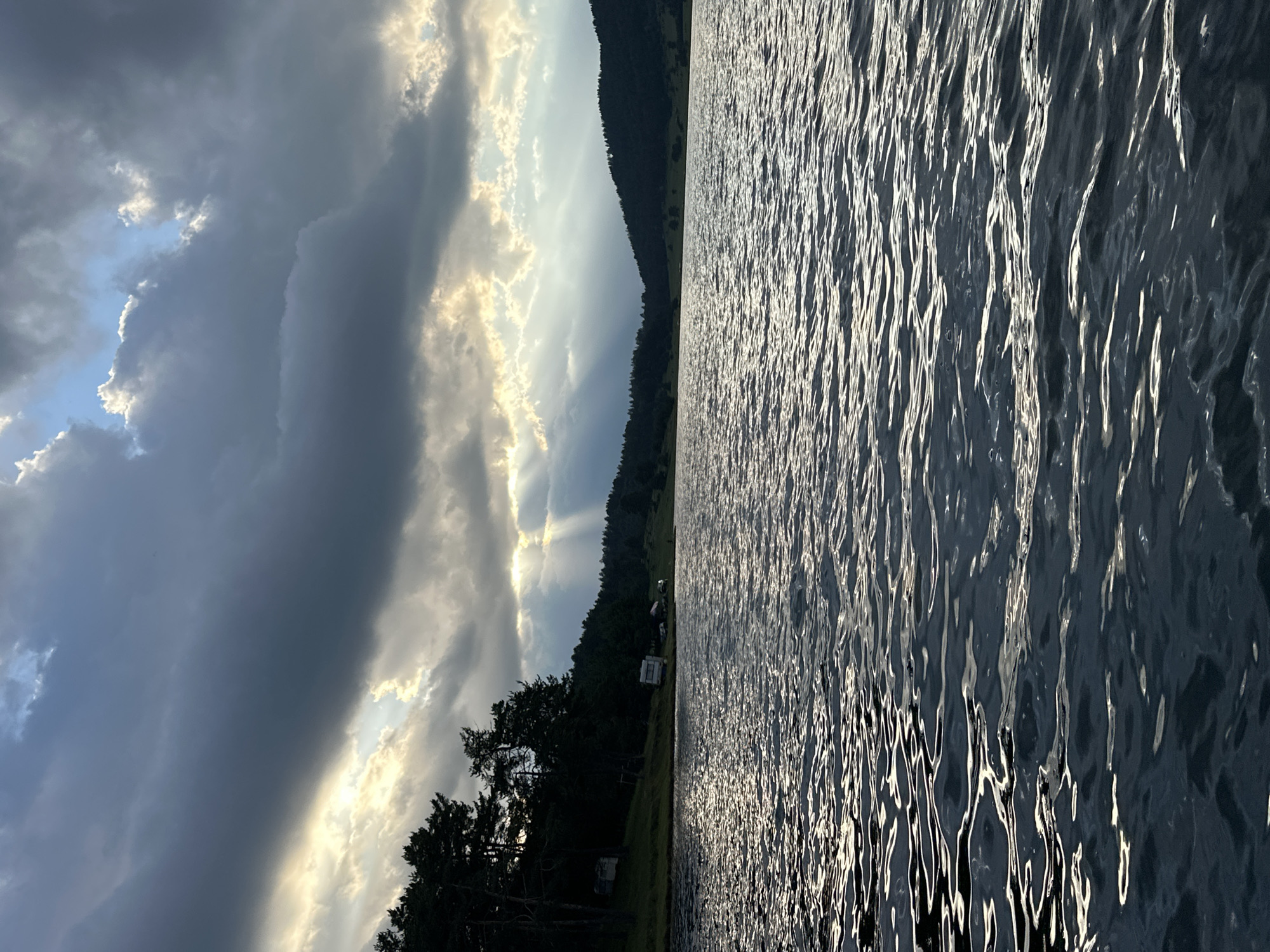 The image depicts a serene lake with choppy water reflecting the dramatic sky. In the background, a tree-covered hillside forms the backdrop, with a few structures, possibly campers or small cabins, nestled amongst the trees. Sunbeams pierce through a dark, cloudy sky, illuminating parts of the water and the distant landscape. The scene suggests a natural, possibly somewhat remote, location.